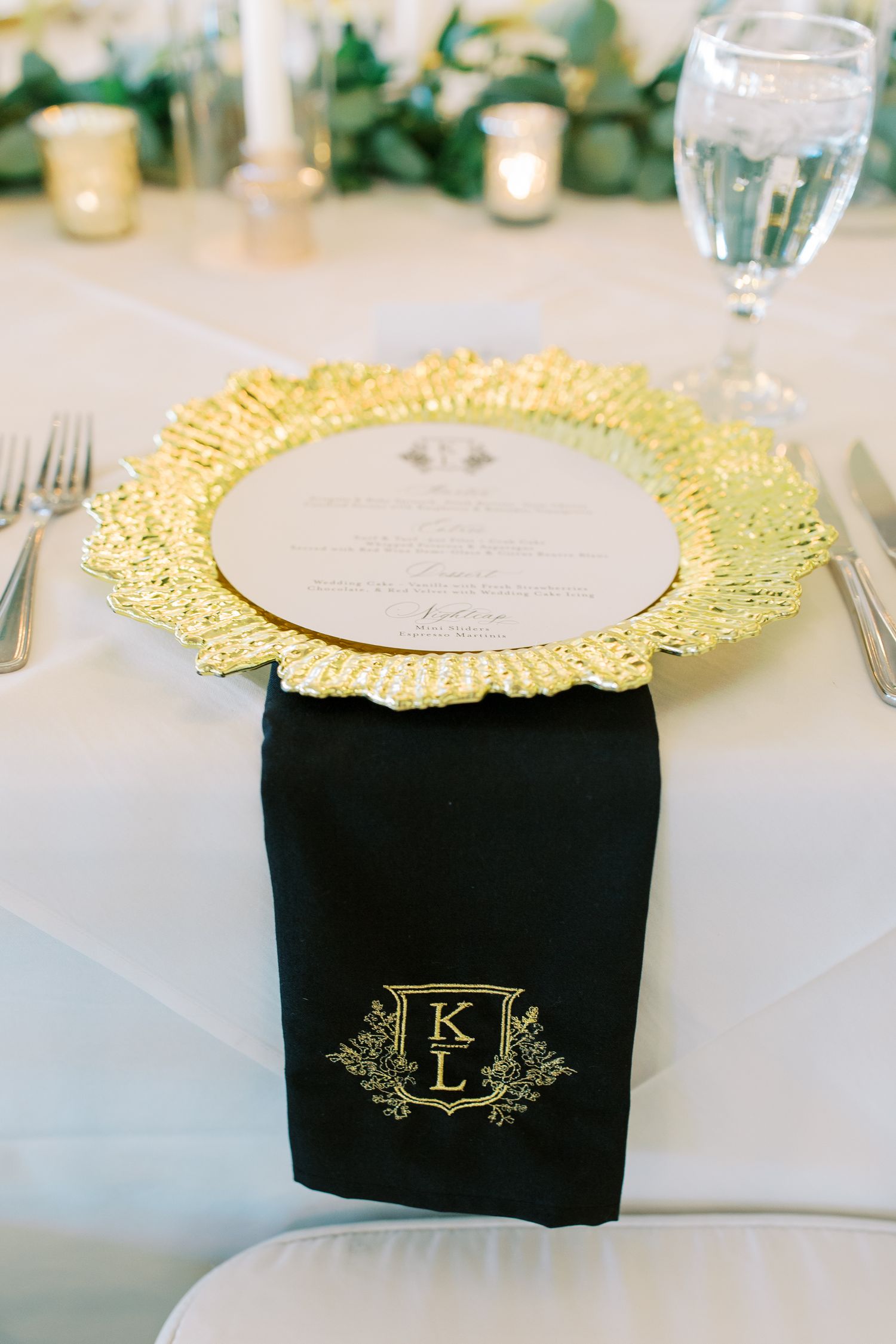 Elegant place setting with gold charger plate and monogrammed black napkin on white tablecloth.