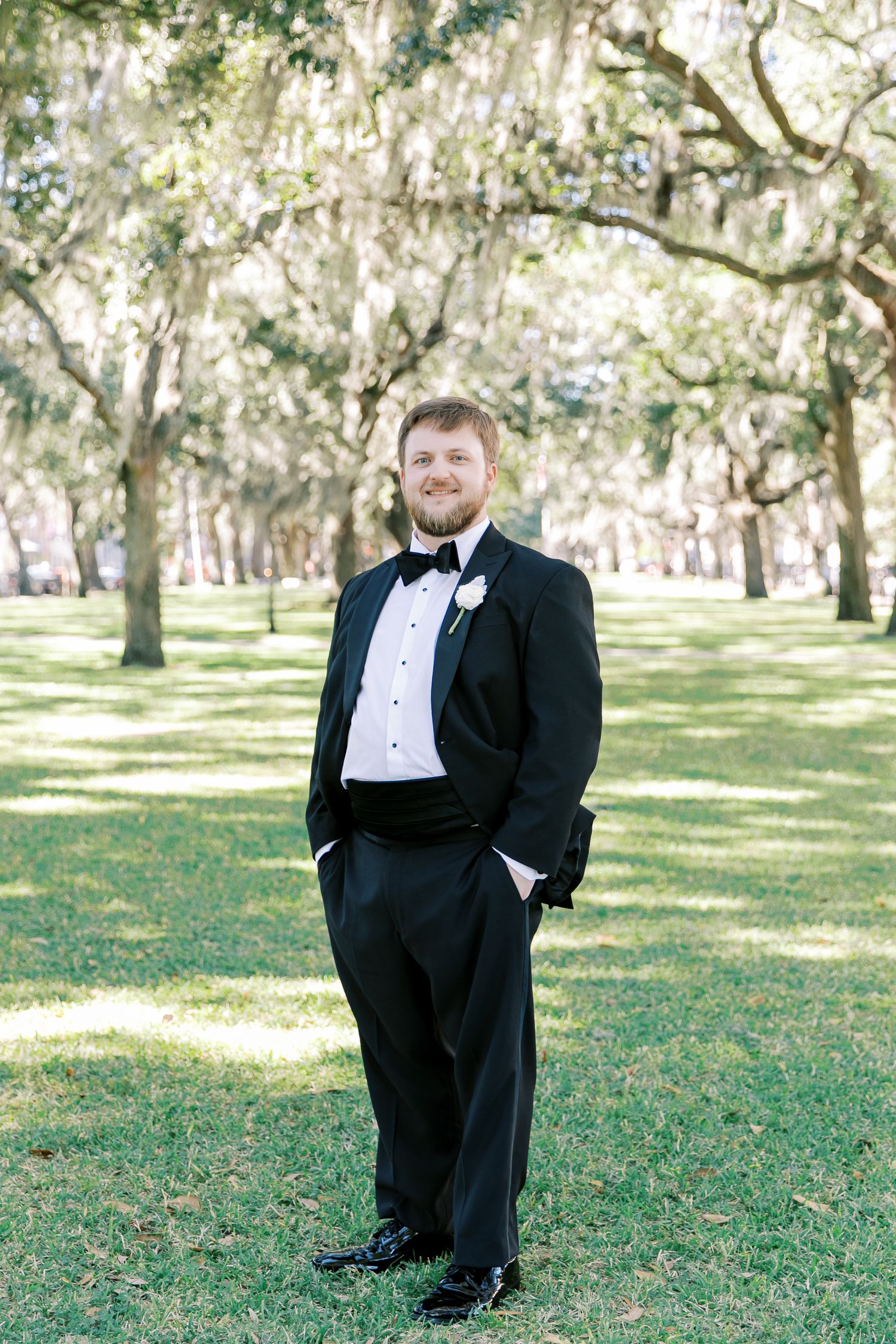 A groom in a classic black tuxedo poses confidently under Spanish moss-draped trees in an outdoor garden setting.