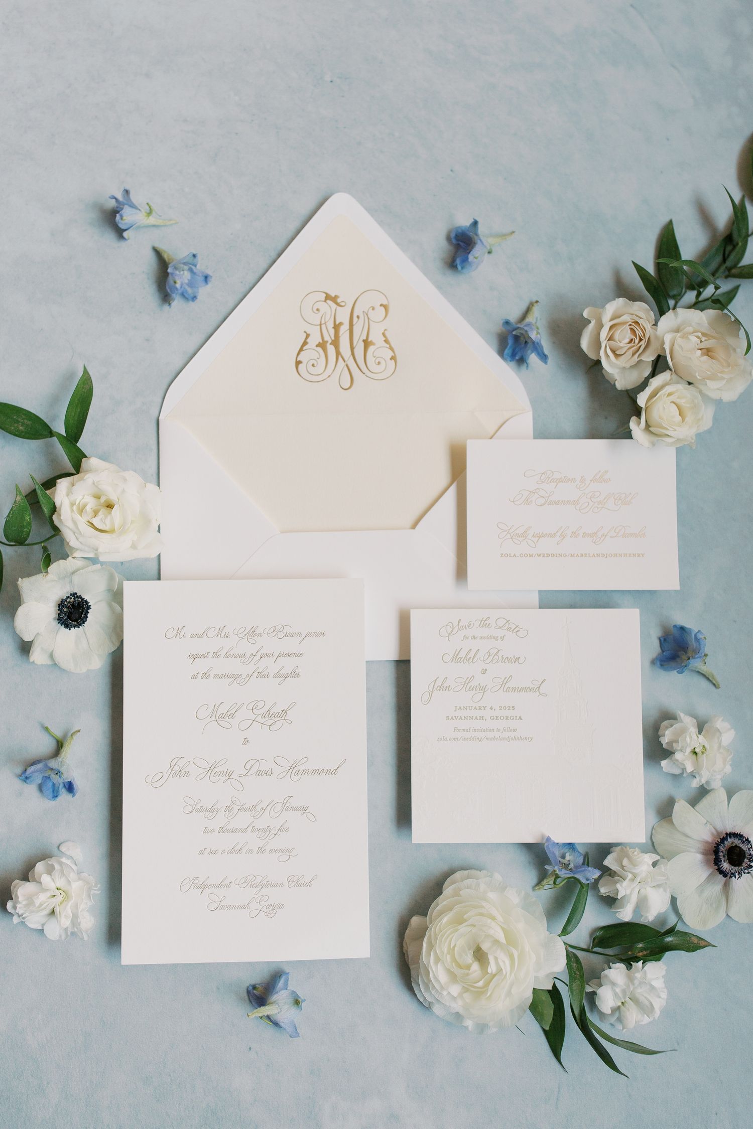 Elegant wedding invitation suite with white envelopes, gold monogram, and blue and white floral accents on gray background.