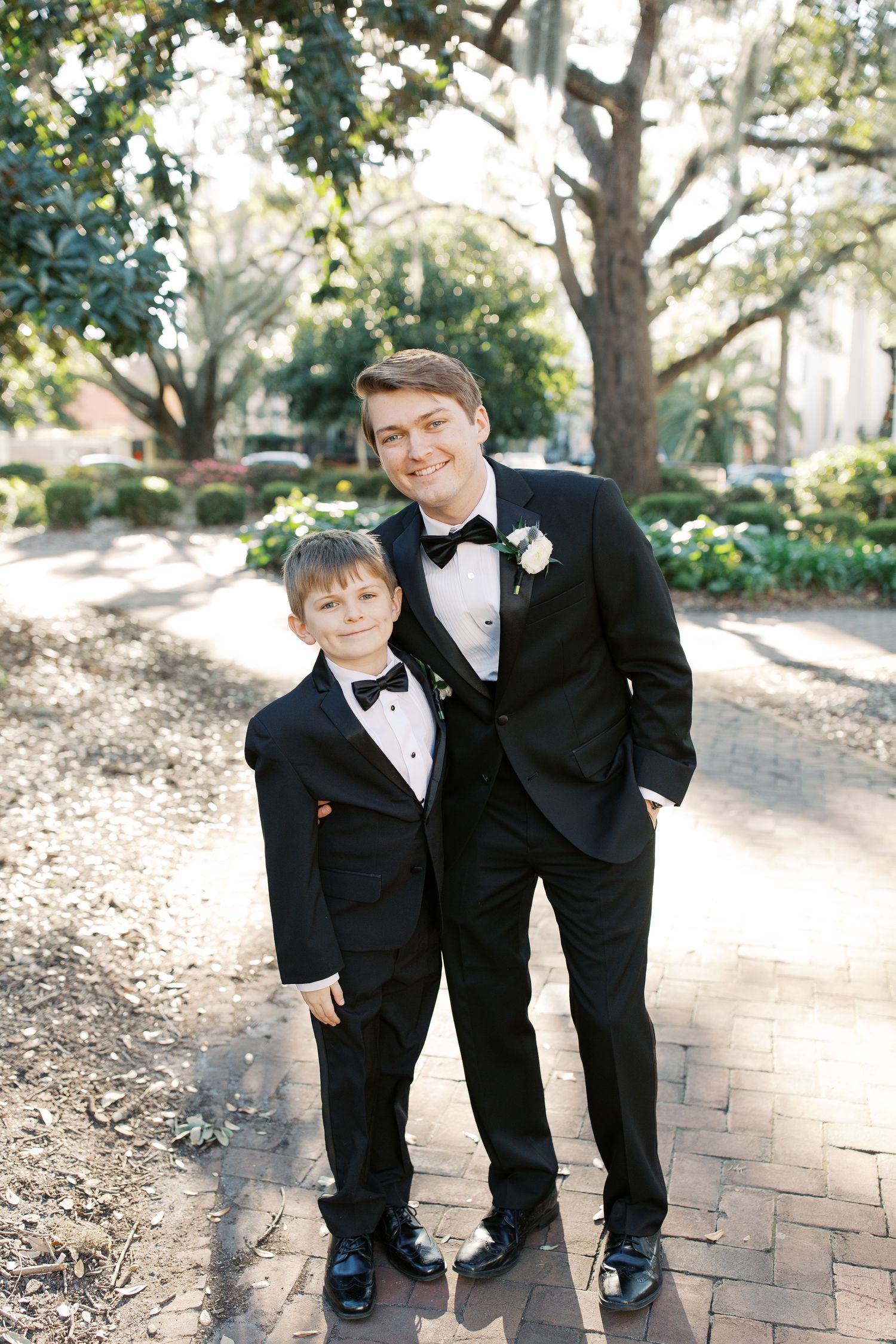 A matching formal pair in black tuxedos pose on a brick pathway under trees.