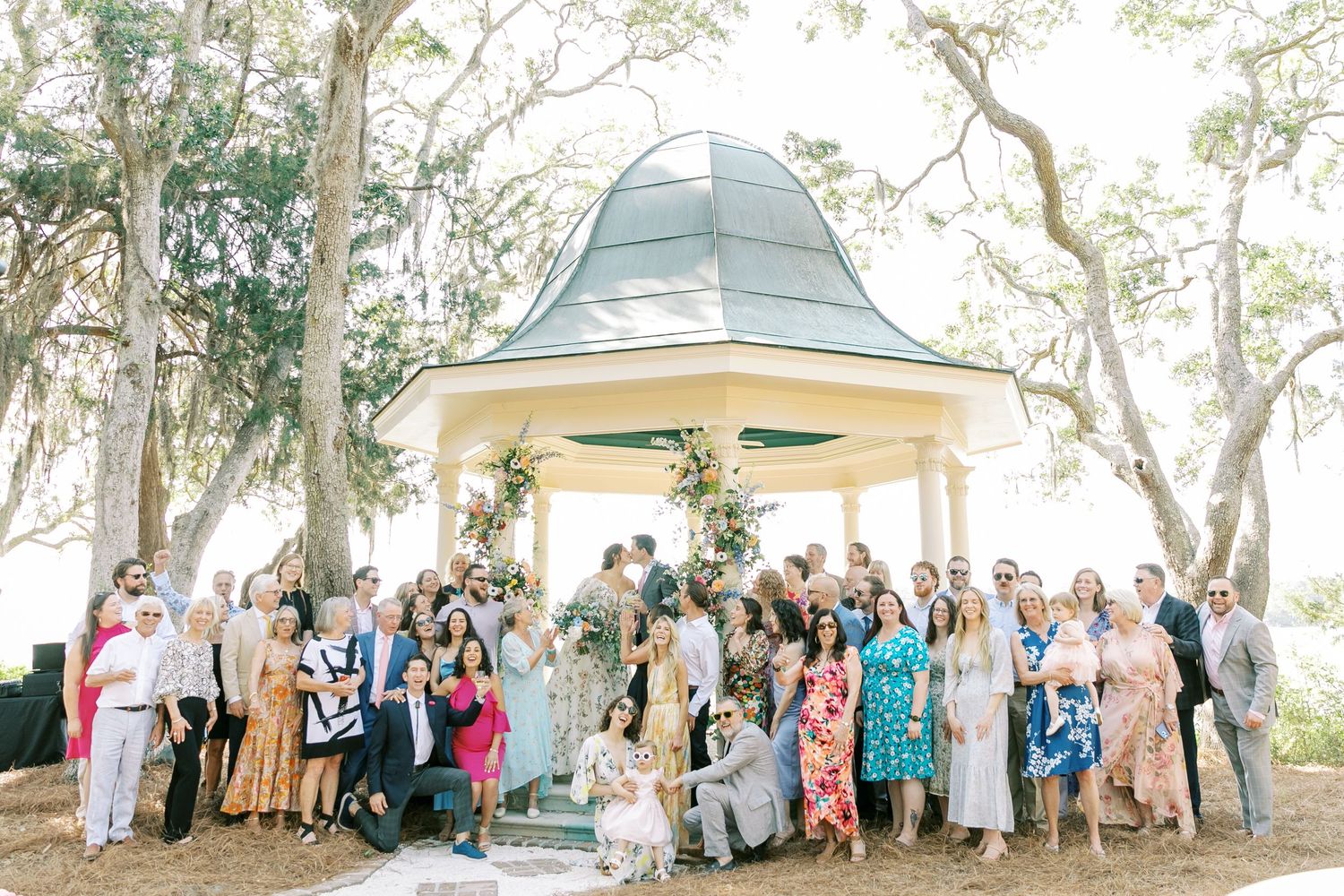 Large group photo of wedding guests gathered under outdoor pavilion surrounded by trees.