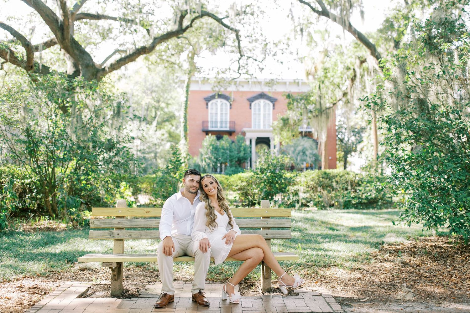 A couple relaxes together on a stone bench in a lush garden setting with a historic brick building in the background.