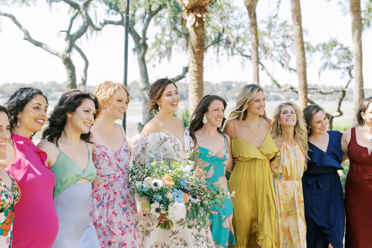 A joyful wedding party poses outdoors in colorful dresses with yellow, pink, and teal tones in a line formation.