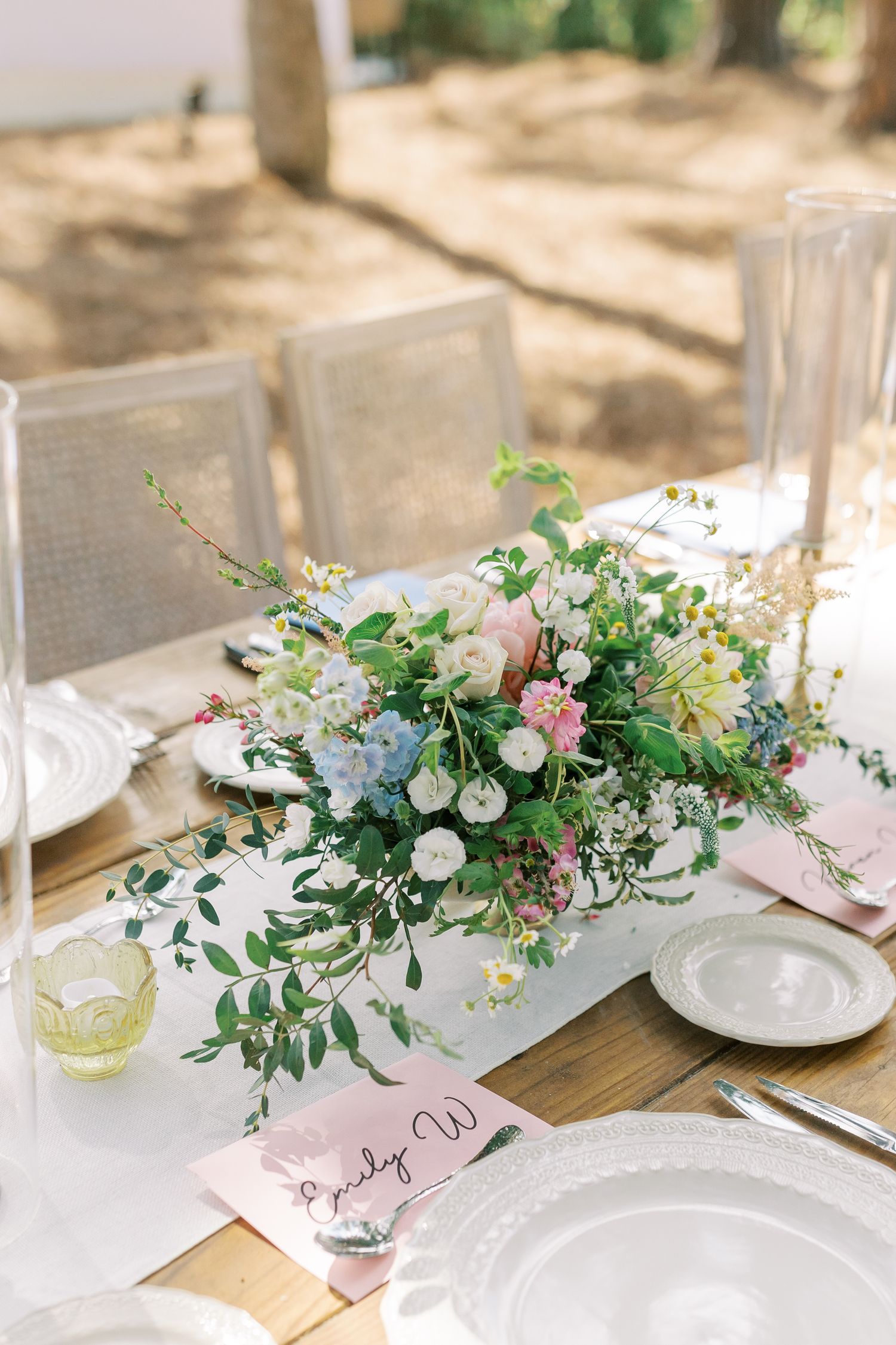 Delicate floral centerpieces with pink and white blooms adorn wooden tables at an outdoor reception.