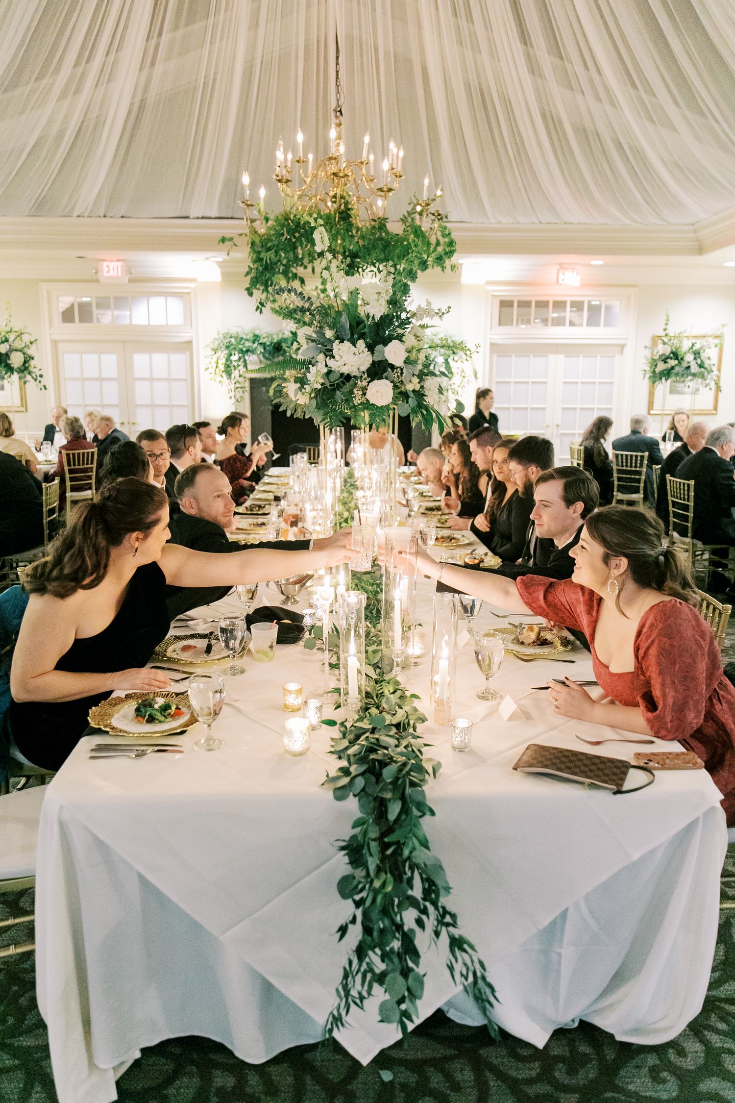 Elegant wedding reception table with draped ceiling, chandelier, and lush greenery garland centerpiece running down white tablecloth.