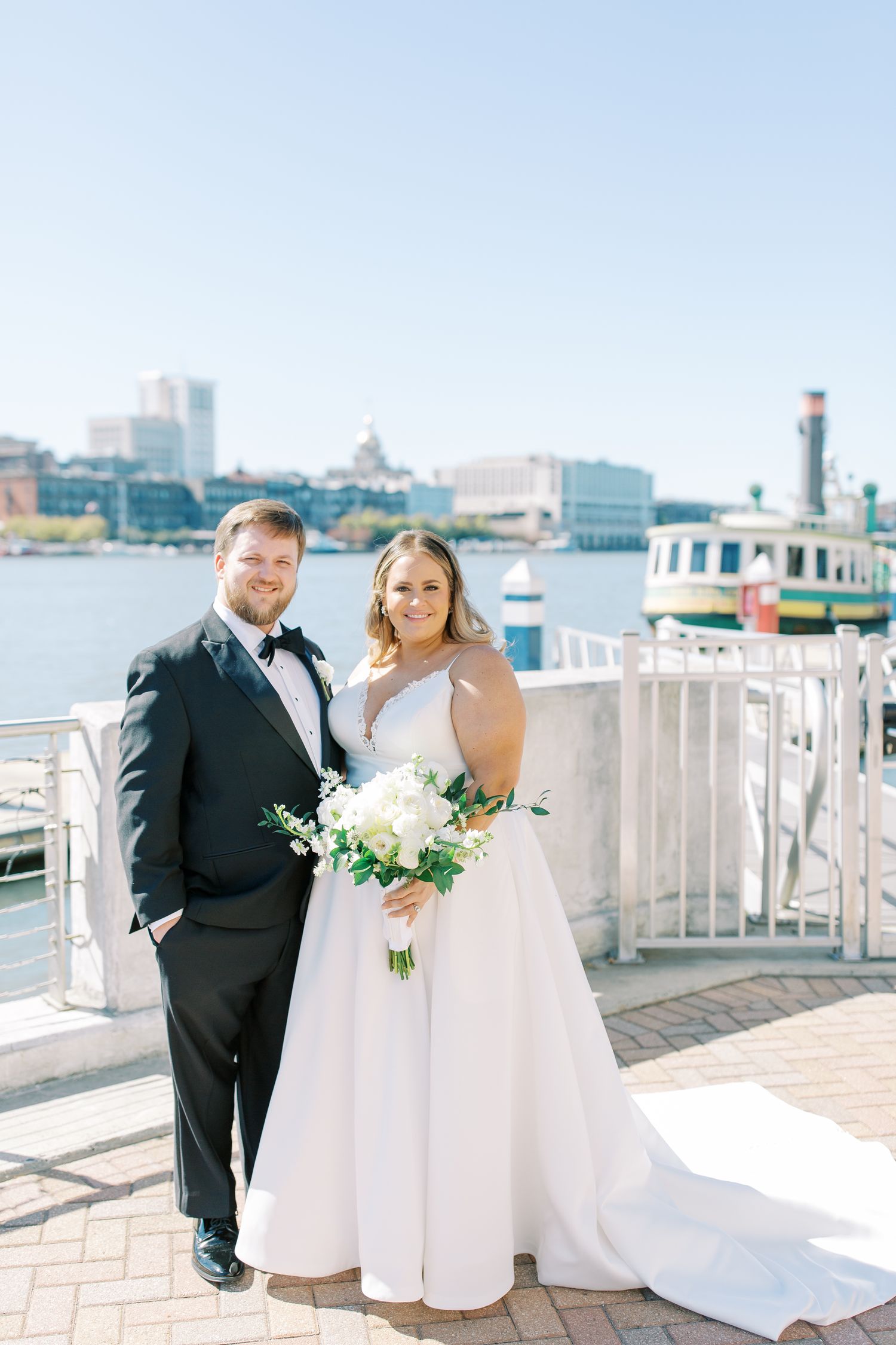 A couple poses on a waterfront deck with a cityscape and harbor in the background.