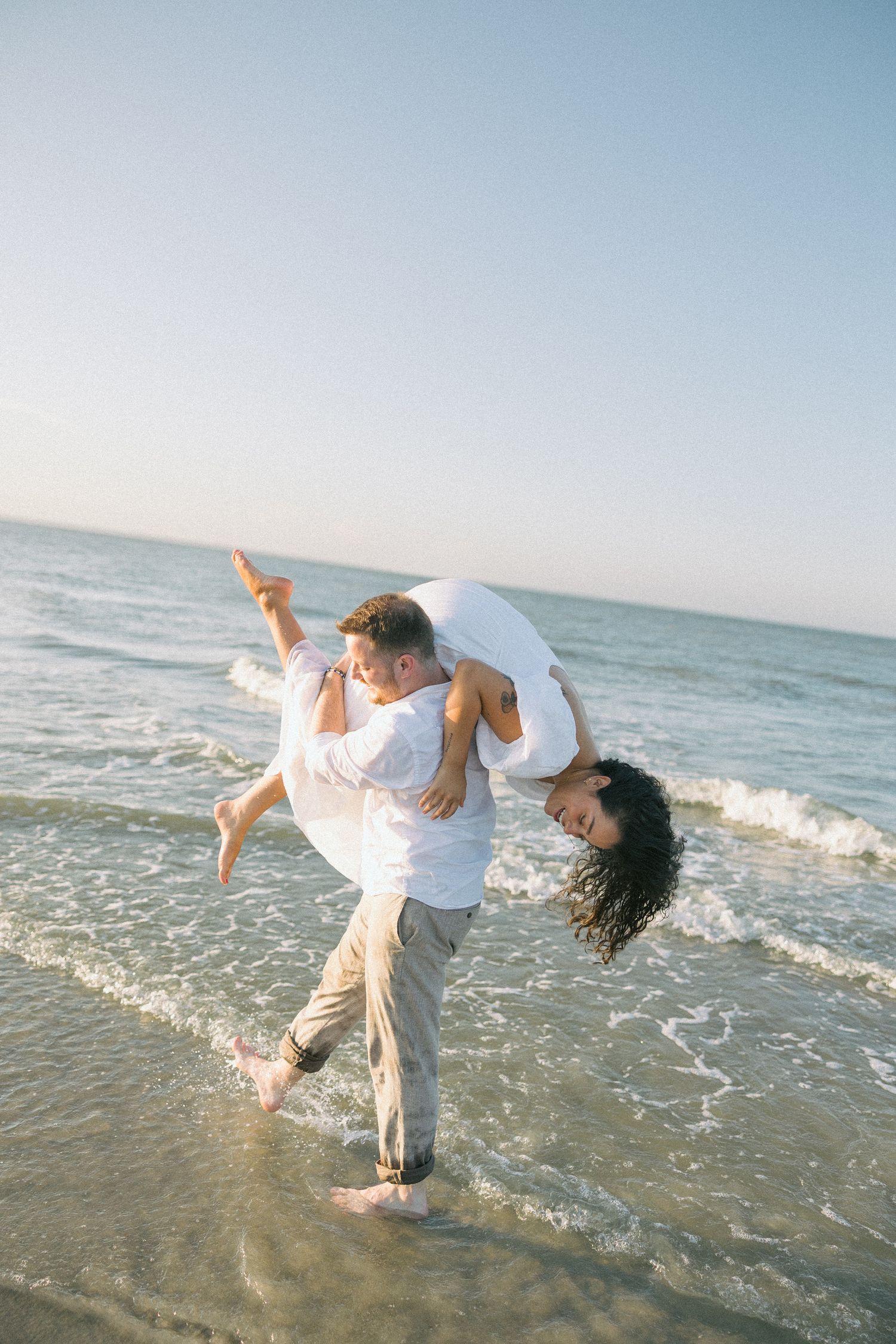A couple dances and splashes in the ocean waves while wearing white clothing.