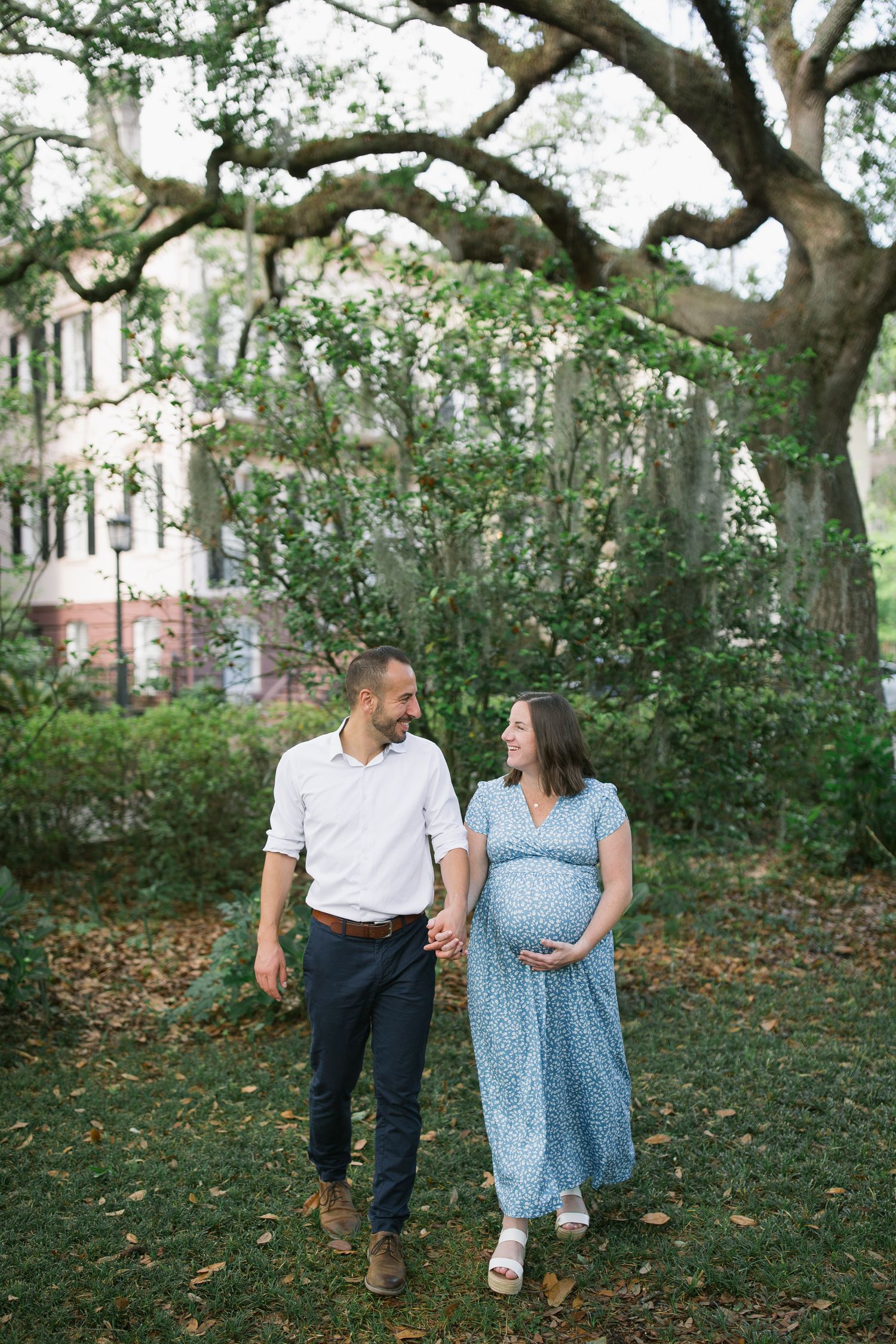 A couple strolling together through a park with large trees and fallen leaves on the ground.