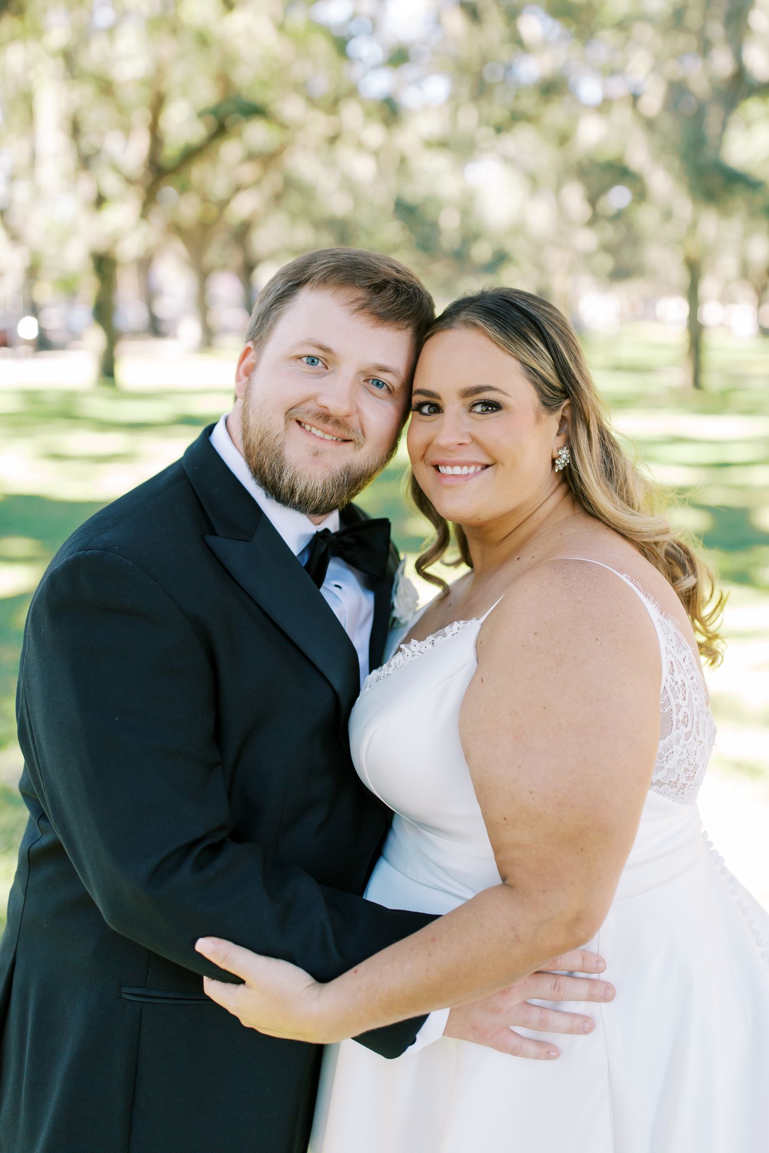 A romantic wedding portrait of a couple in formal attire smiling together in a sunny outdoor garden setting.
