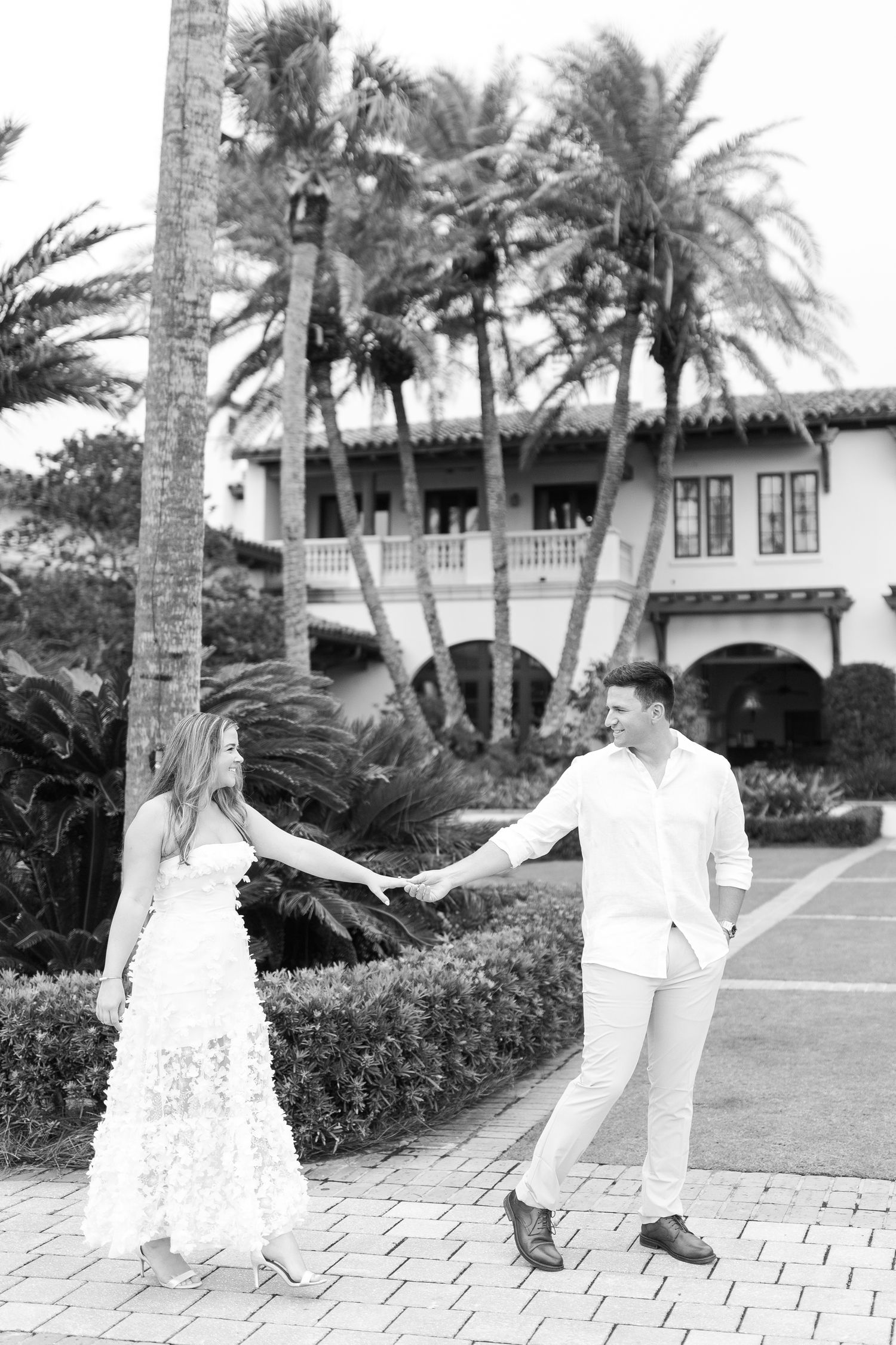 Black and white photo of couple dancing on paved walkway with palm trees and Spanish architecture in Sea Island, GA