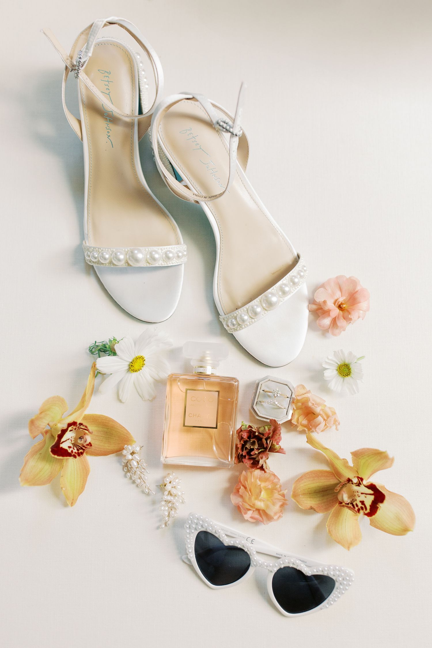 Wedding accessories including white pearl sandals, sunglasses, perfume and peach flowers on a light background.
