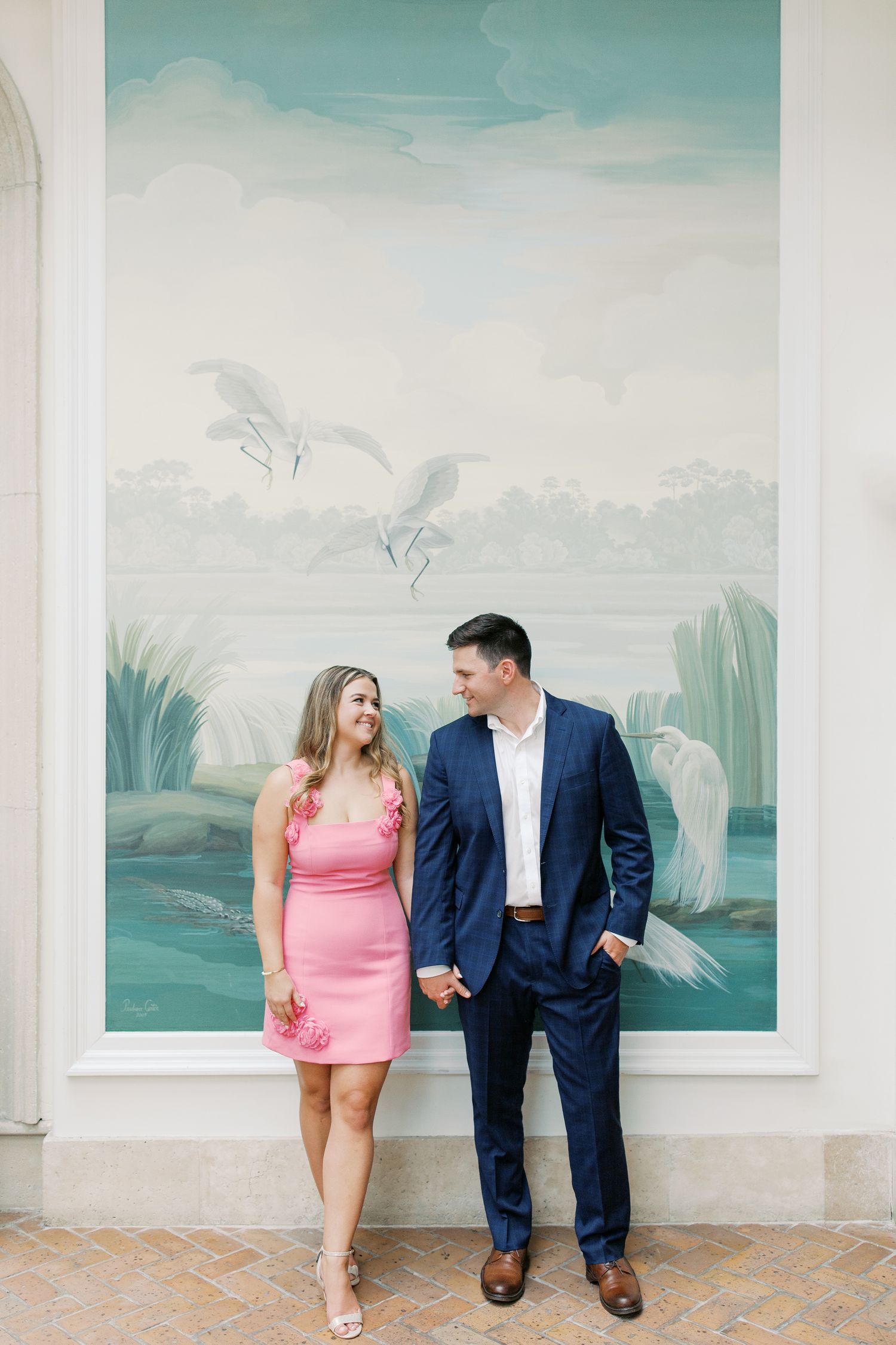 A couple stands in front of a pale mint green wall mural, dressed in a pink dress and navy suit in Sea Island, GA