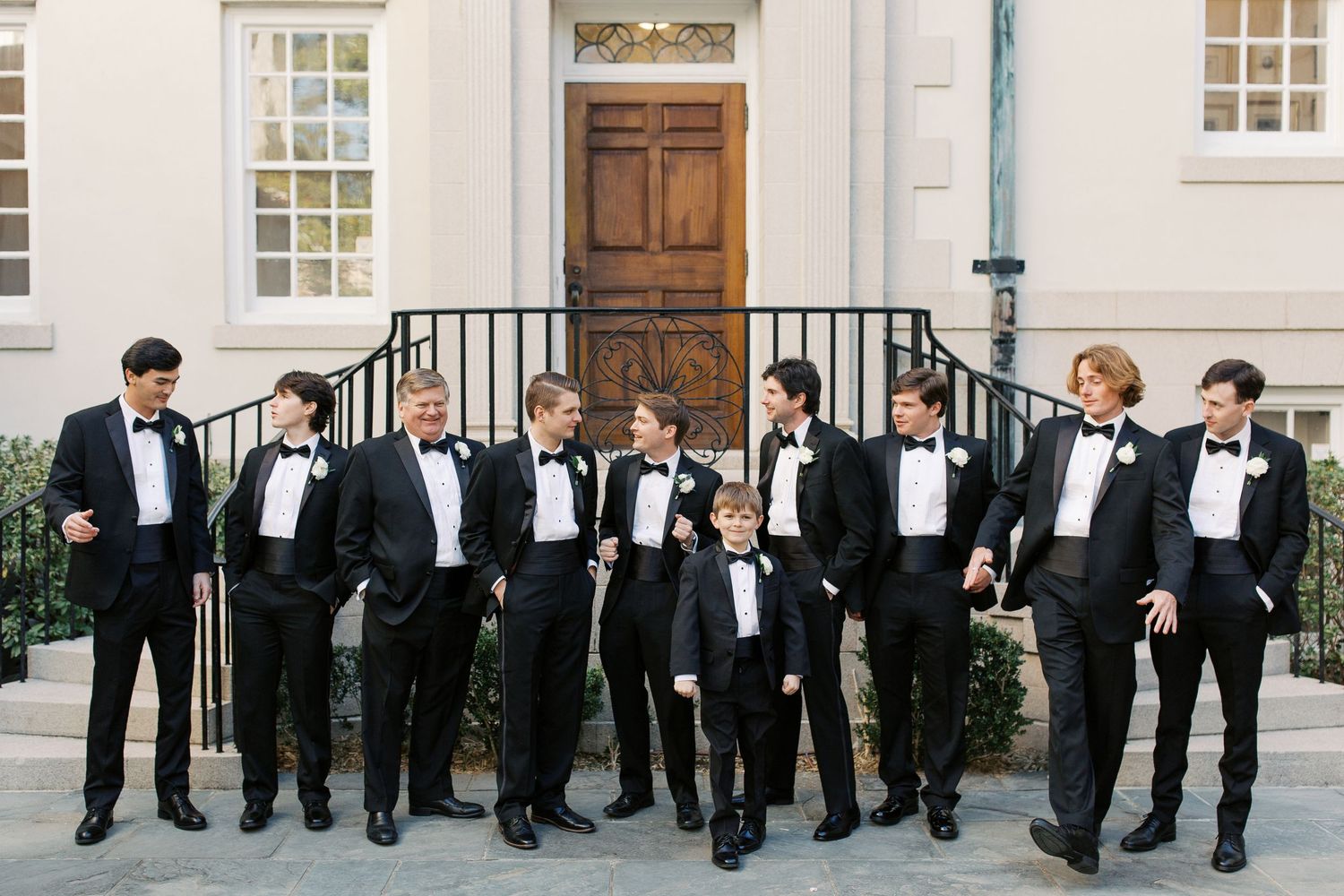 Large group of groomsmen in formal black tuxedos posing outside traditional building.