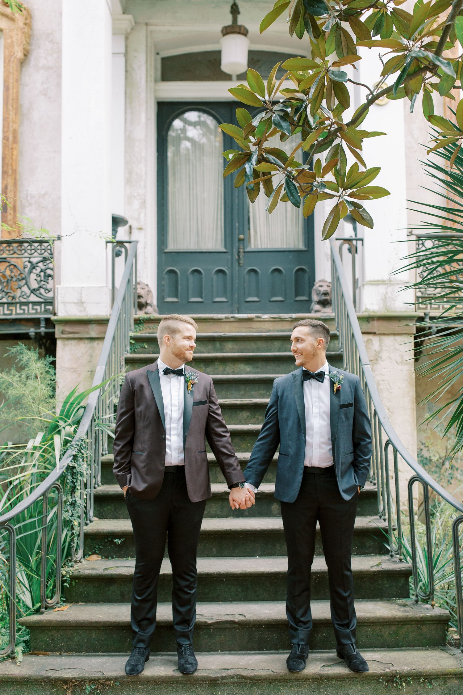 A gay couple in matching black tuxedos and bow ties hold hands while standing on the steps of a historic southern home.