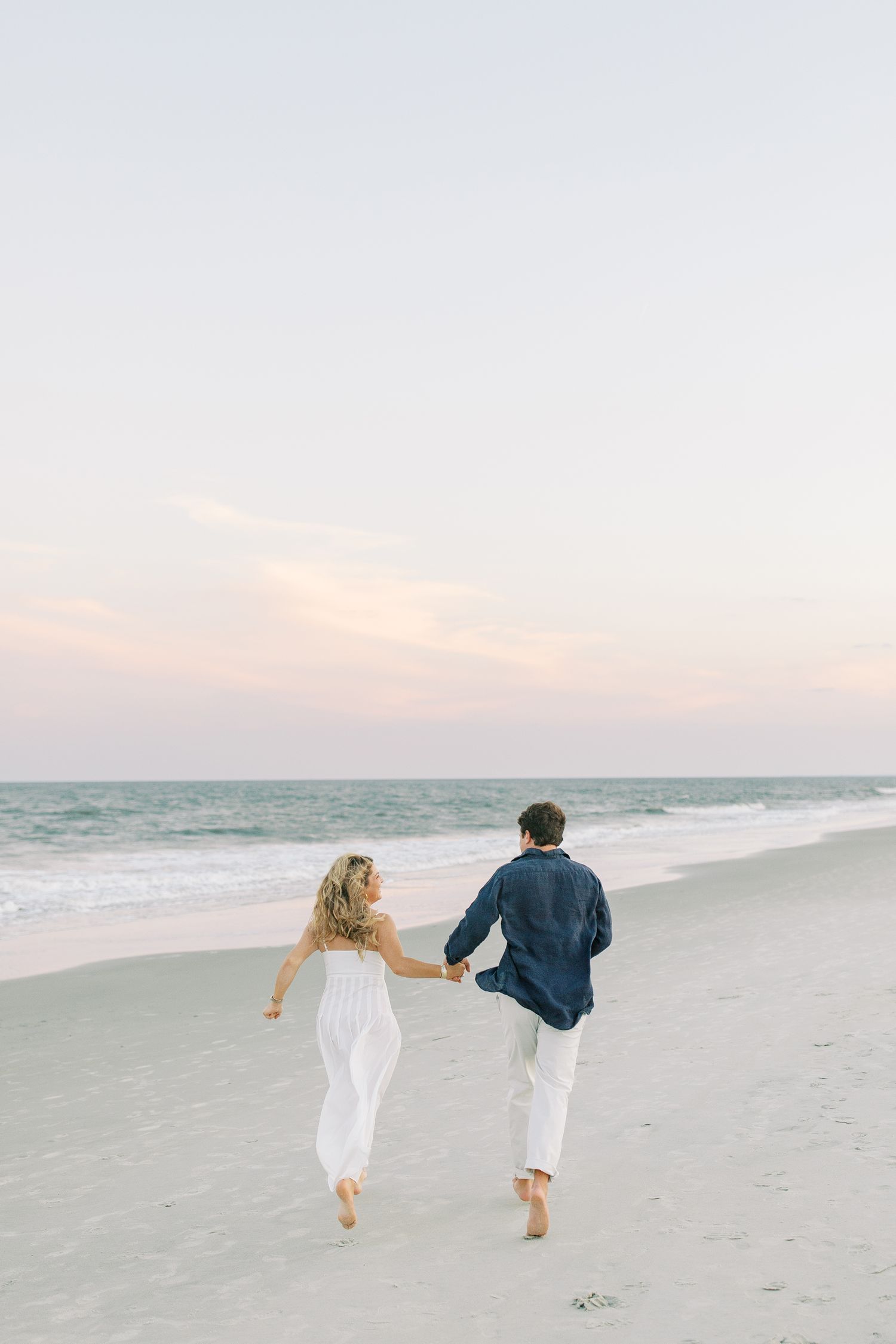 A couple runs barefoot along a peaceful beach at sunset, with the ocean waves gently rolling in beside them.