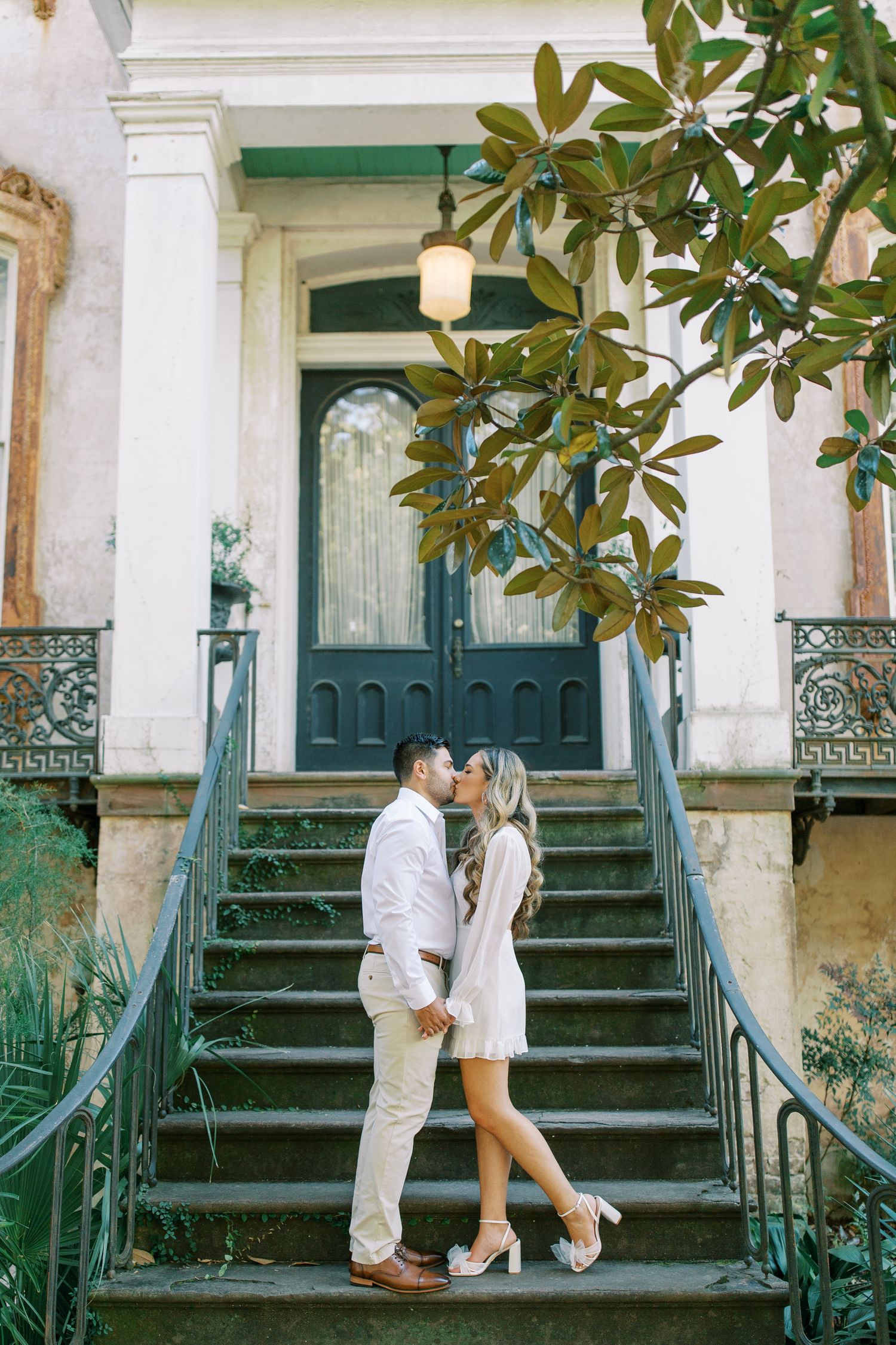 Couple sharing a romantic moment on vintage green wrought iron stairs leading to an ornate doorway with magnolia tree.