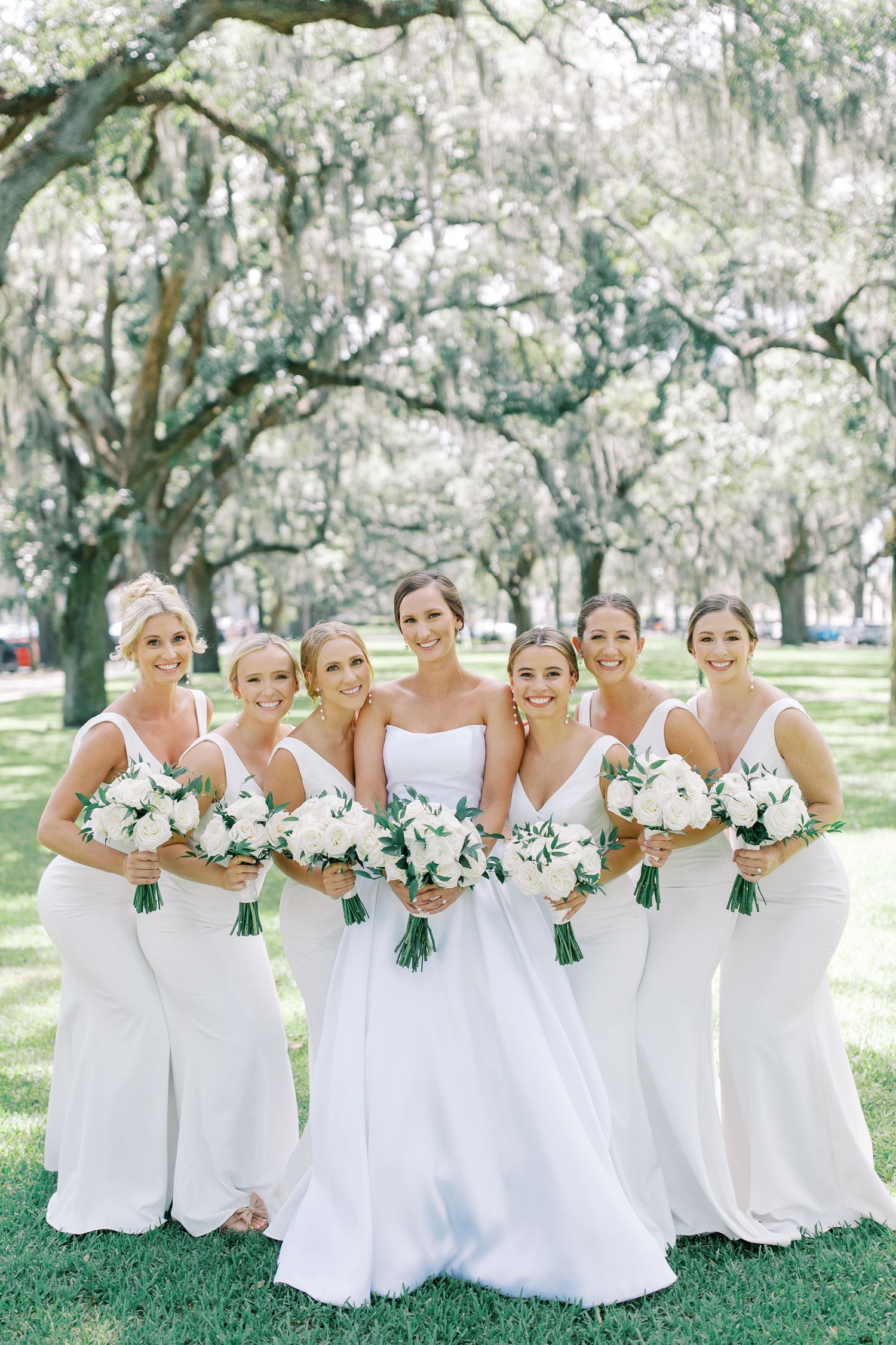 Bridesmaids in elegant white dresses gather under Spanish moss draped trees holding white floral bouquets.