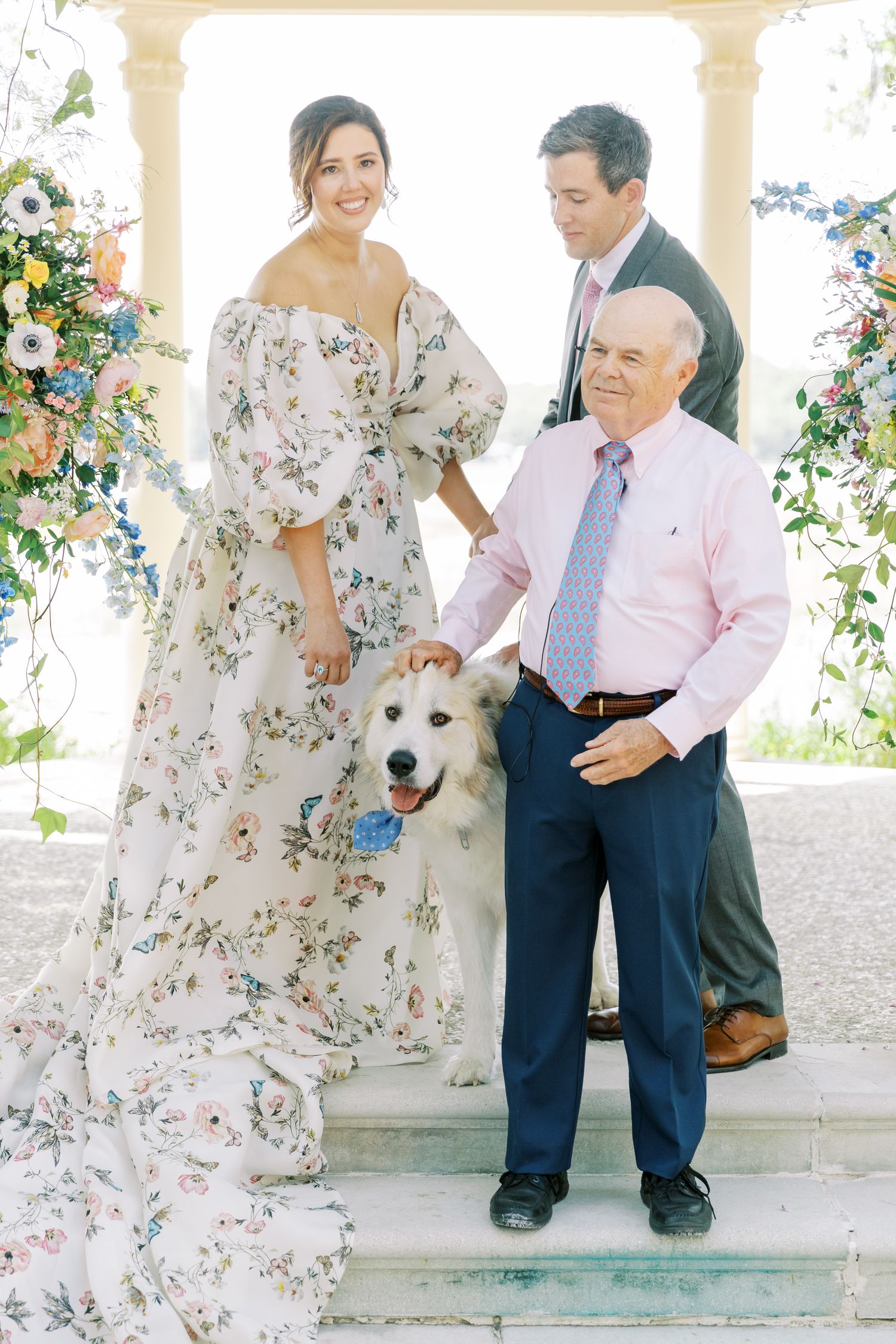 A white dog sits outdoors with people on stone steps during a sunny celebration.