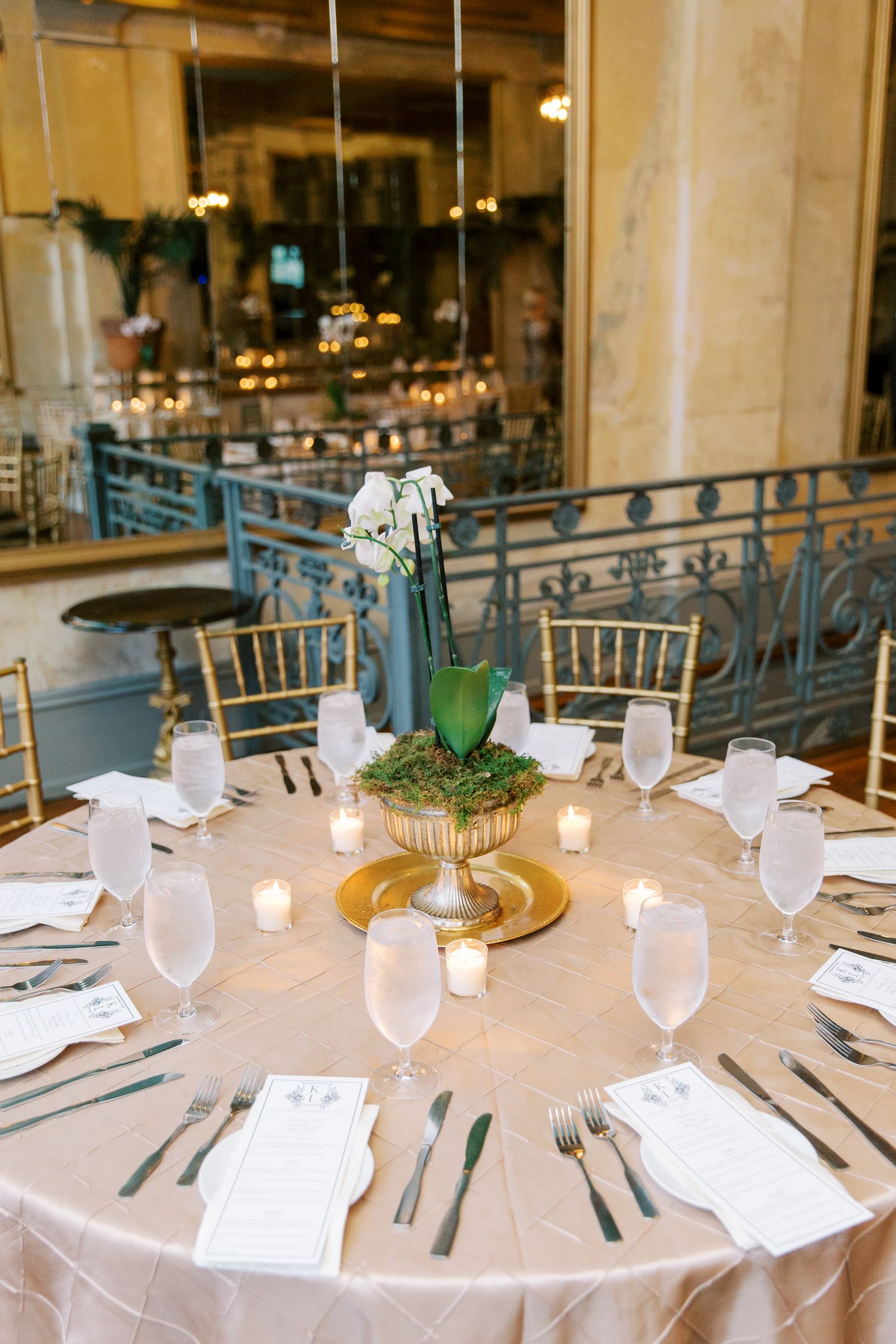 An elegant round wedding reception table set with frosted glassware, white linens, and a gold centerpiece vase with orchids.