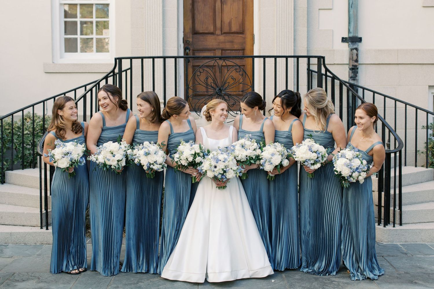 A wedding party in blue bridesmaid dresses and white bridal gown poses on steps of a traditional building.