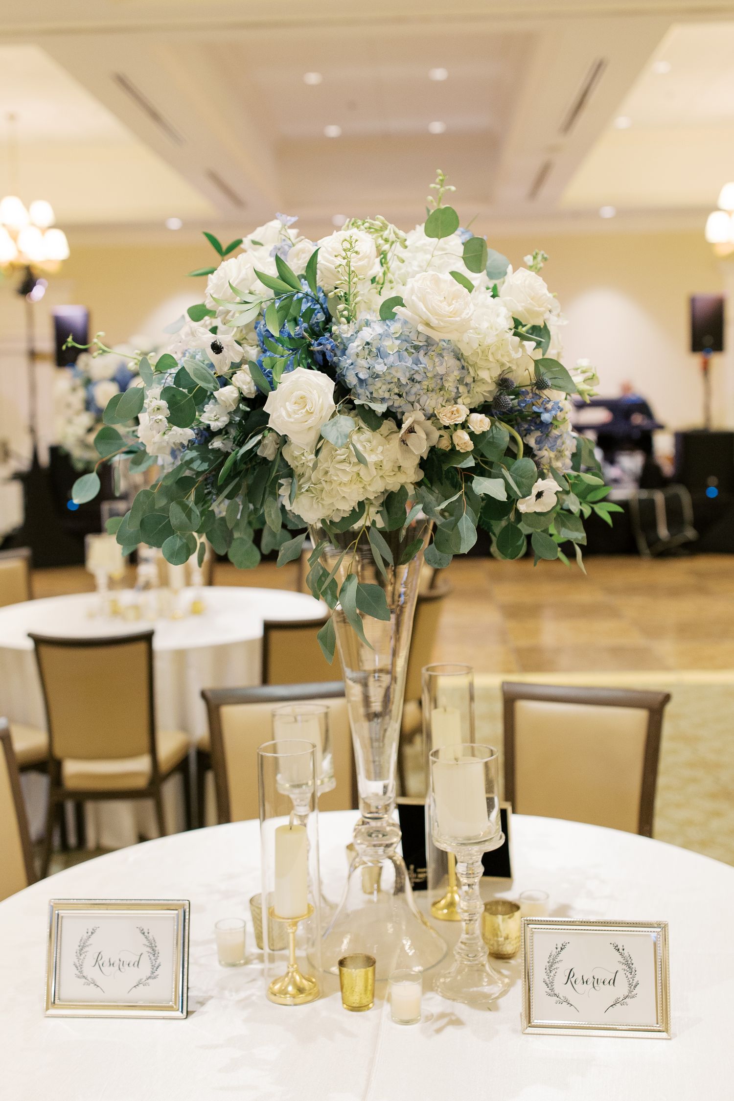 Wedding reception centerpieces with white and blue flowers in tall vases on round tables.