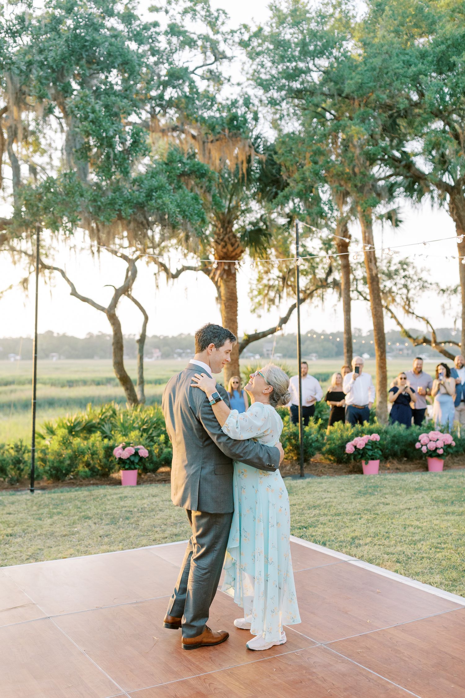 Couple shares a romantic first dance on an outdoor terrace surrounded by oak trees and pink flowers at sunset.