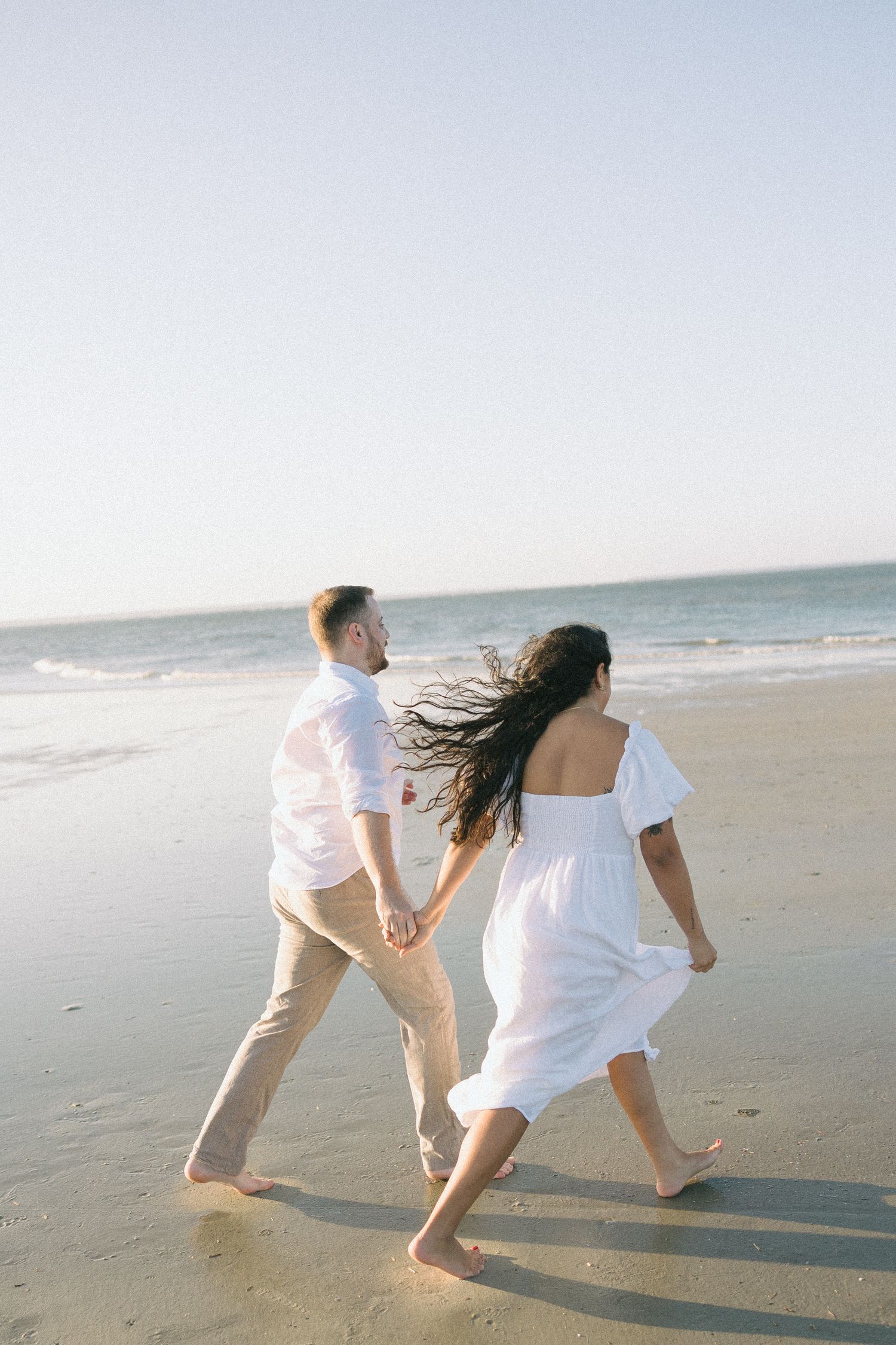 A couple in white clothing runs playfully along a sandy beach at sunset with their hair blowing in the wind.