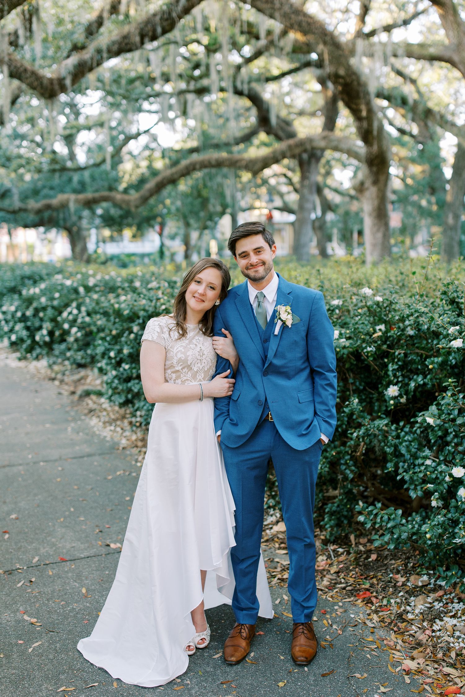 A couple poses under moss-draped oak trees in a garden setting, wearing wedding attire including a blue suit and white gown.
