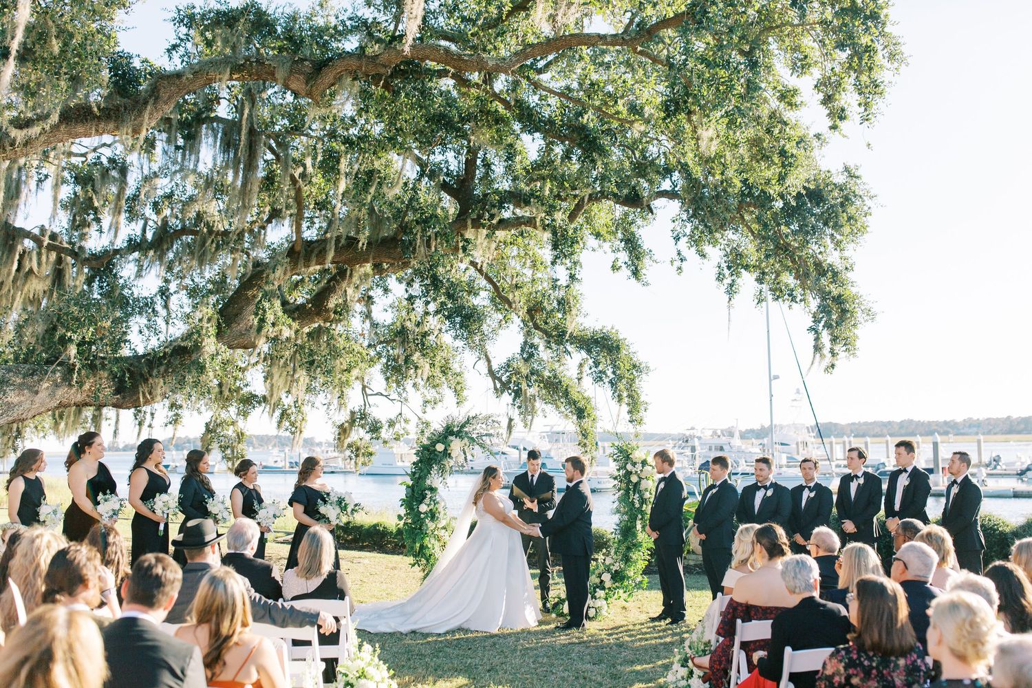 Outdoor wedding ceremony under a large oak tree draped with Spanish moss overlooking the water.