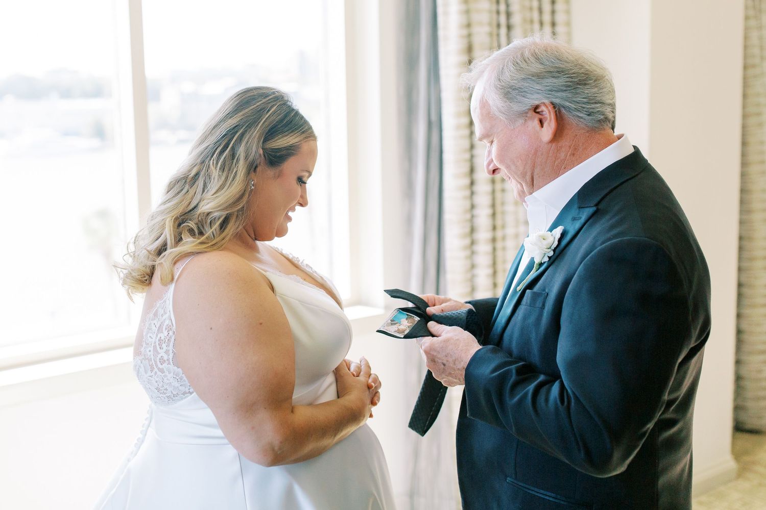Two people in formal attire stand near a bright window having an intimate moment during a special occasion.