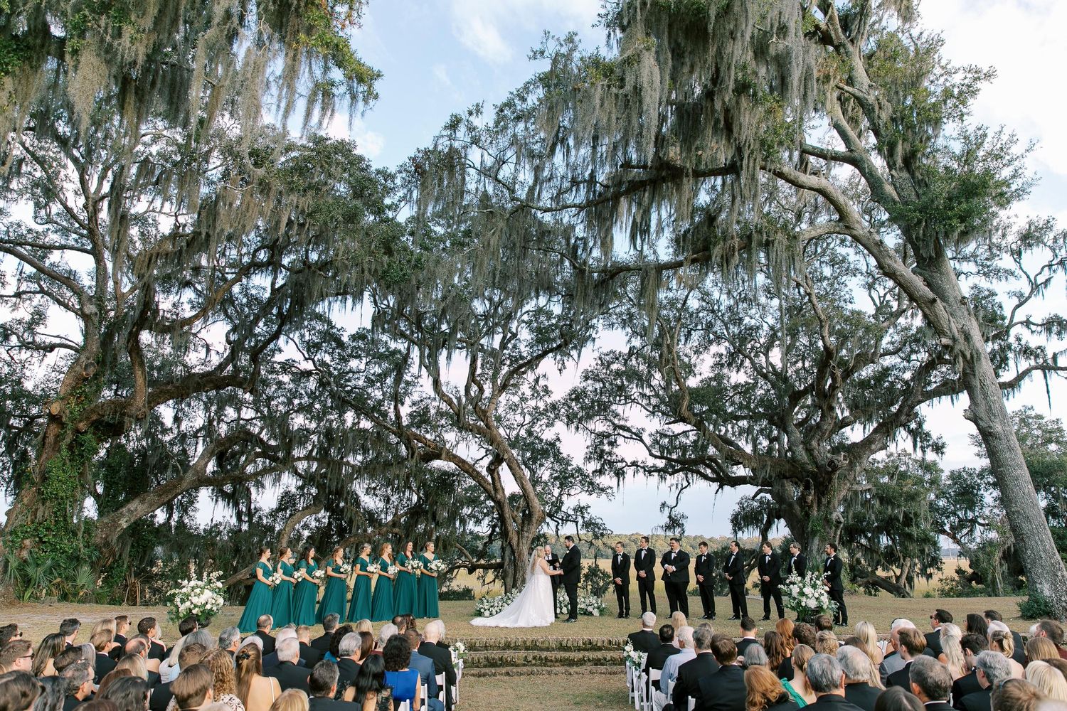 An outdoor wedding ceremony takes place under majestic oak trees draped with Spanish moss as guests watch from their seats.
