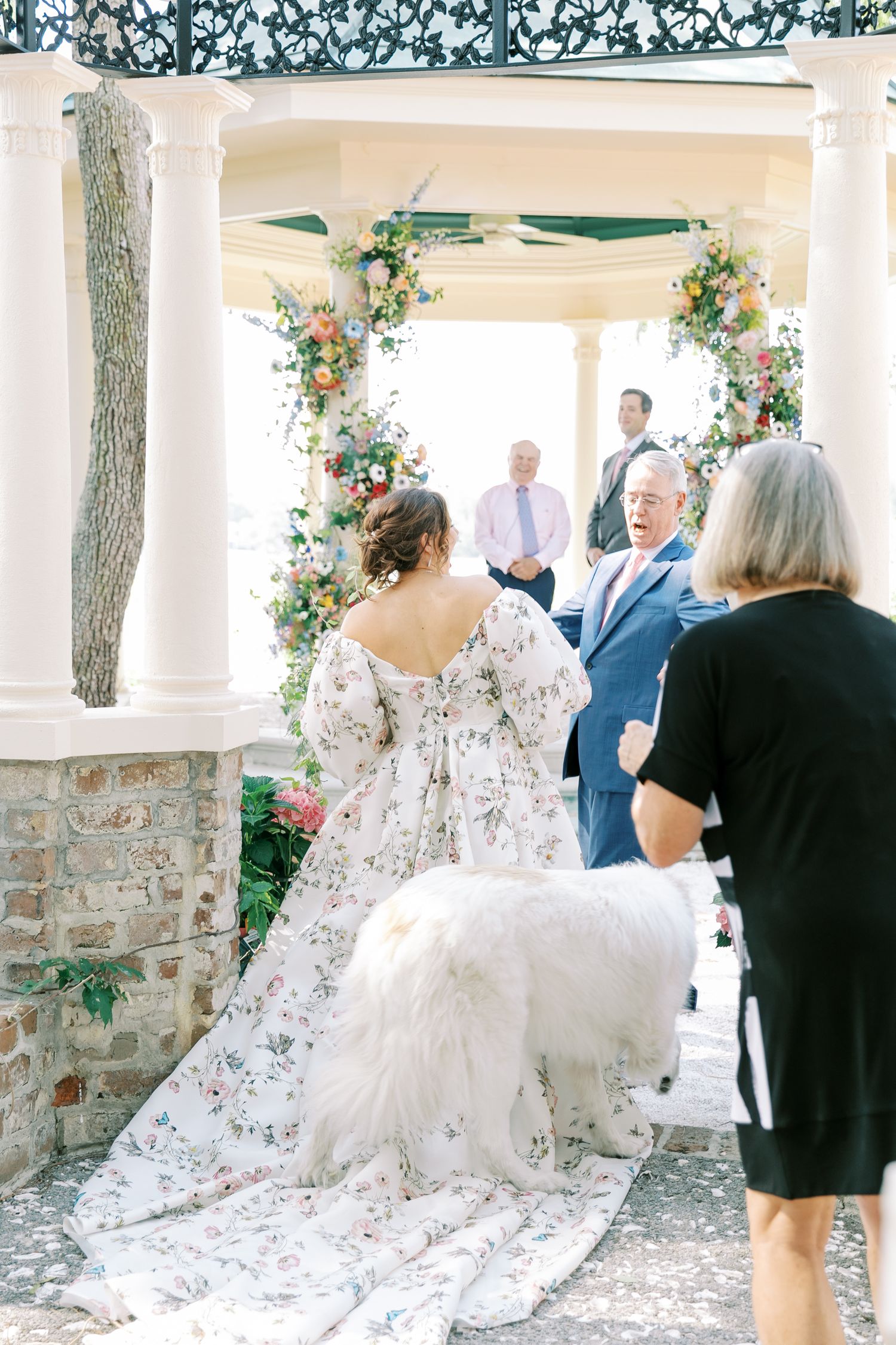 A wedding ceremony in a gazebo decorated with floral arrangements and white columns alongside a stone wall.