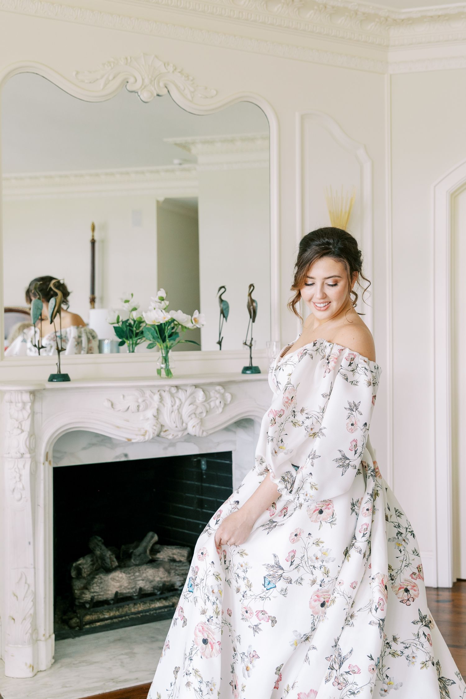Elegant off-shoulder floral gown with long sleeves shown against an ornate white fireplace with classic mirror.