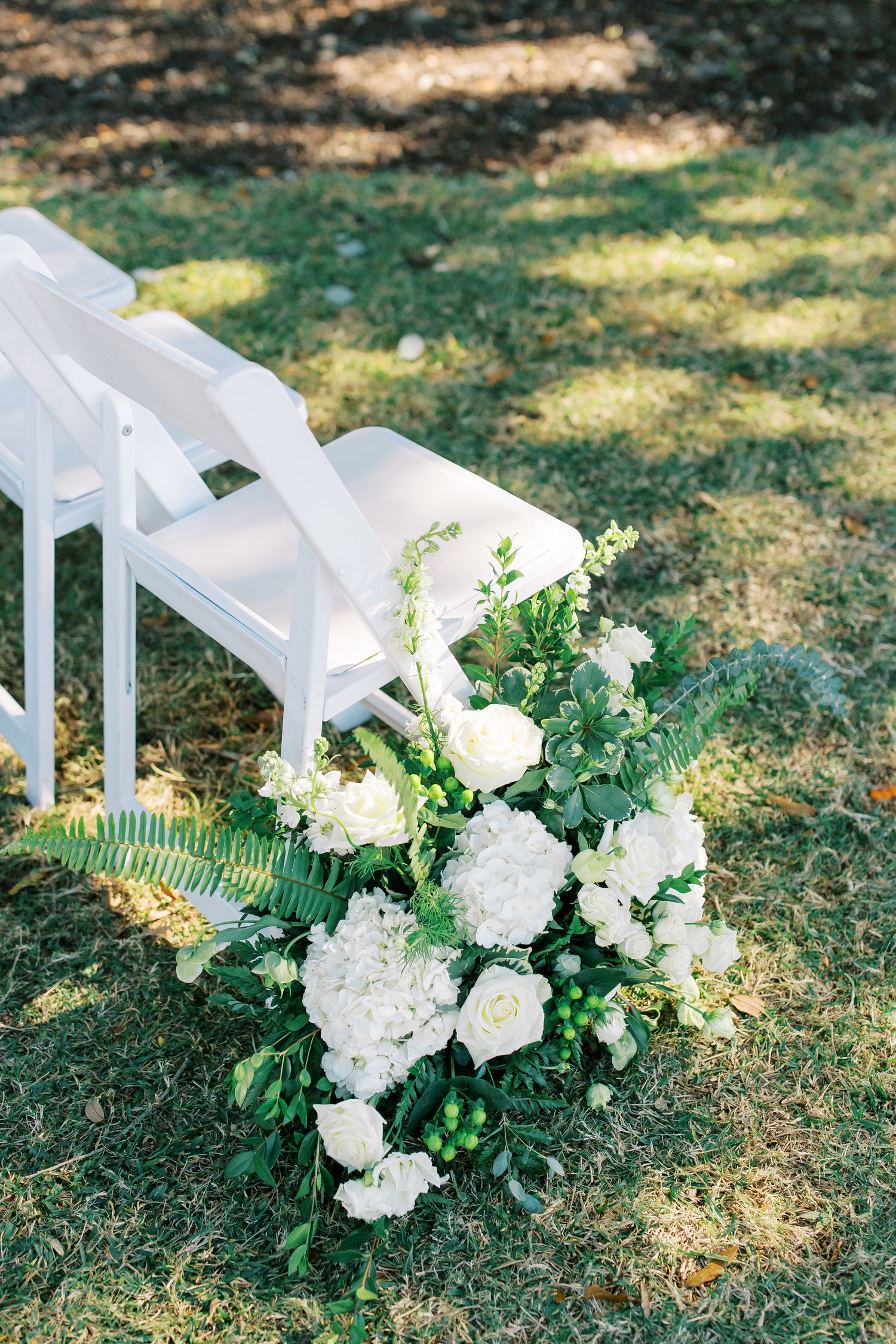White floral arrangement with hydrangeas and greenery decorating white ceremony chairs outdoors.