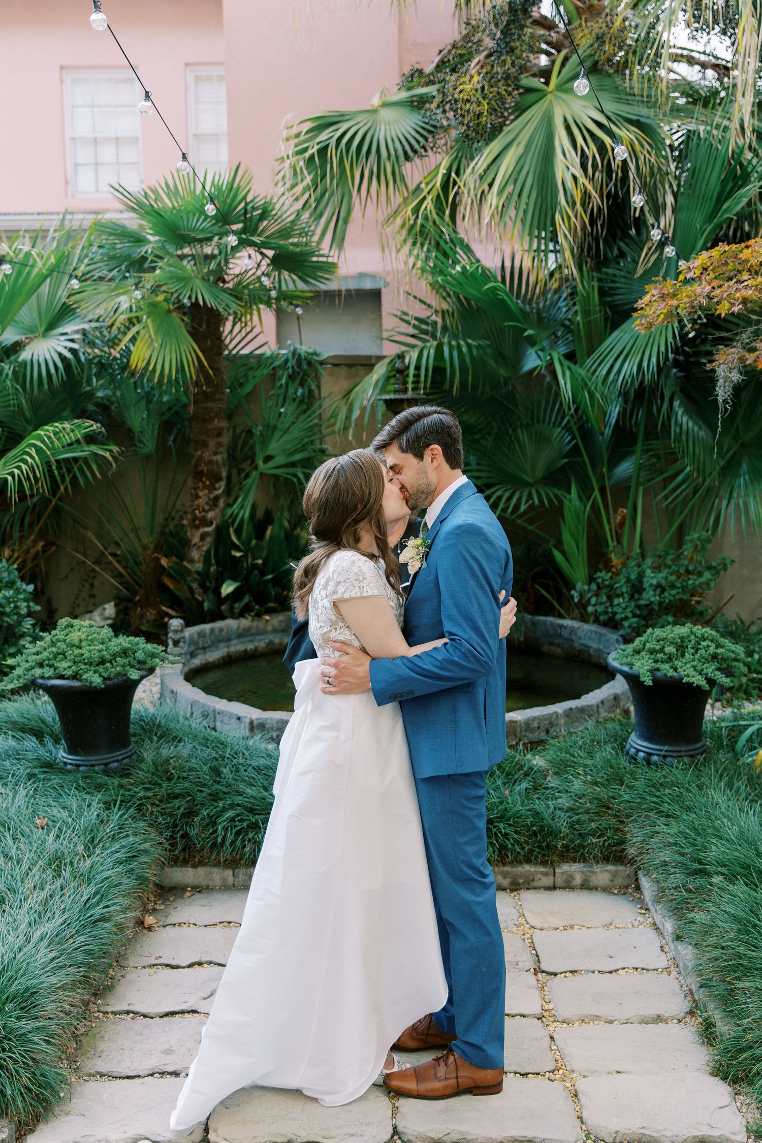 A wedding couple shares tender moments in a lush tropical garden setting with pink walls and palm trees.