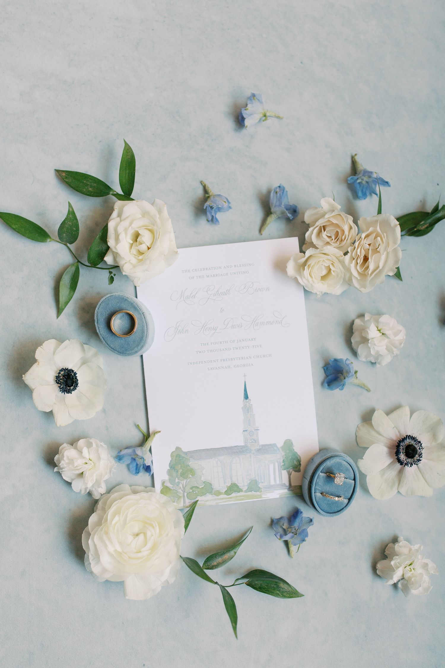 Wedding stationery layout with white roses, blue flowers, and greenery on a light gray background.