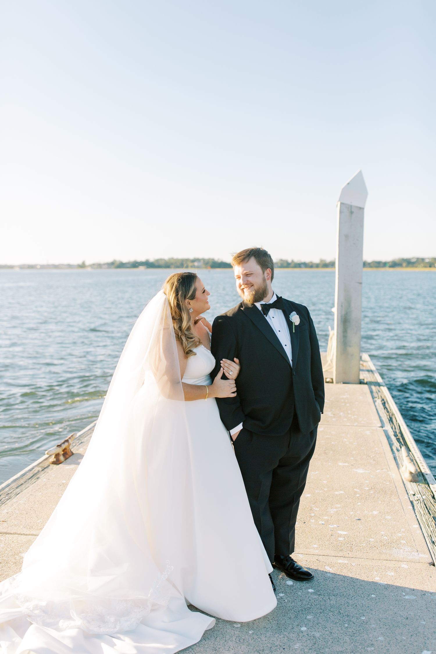 A happy couple in wedding attire share a joyful moment together on a pier overlooking a serene waterfront.