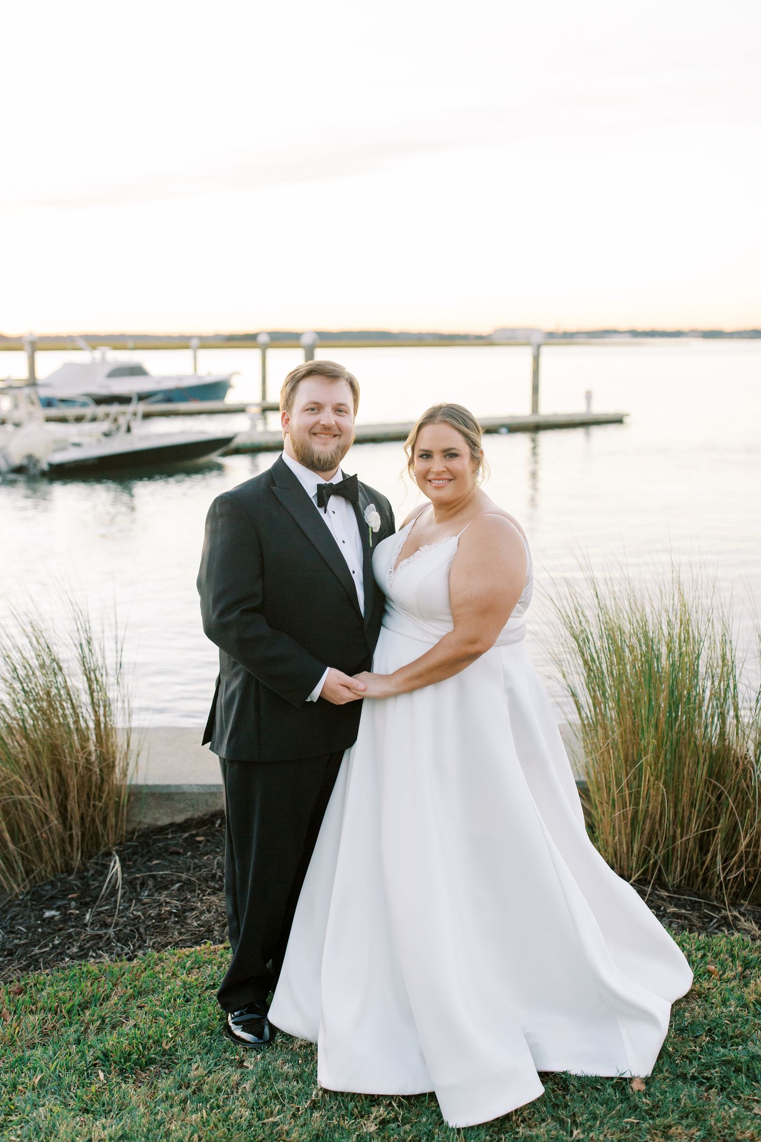 Wedding couple posing by marina with boats and water in background at sunset.