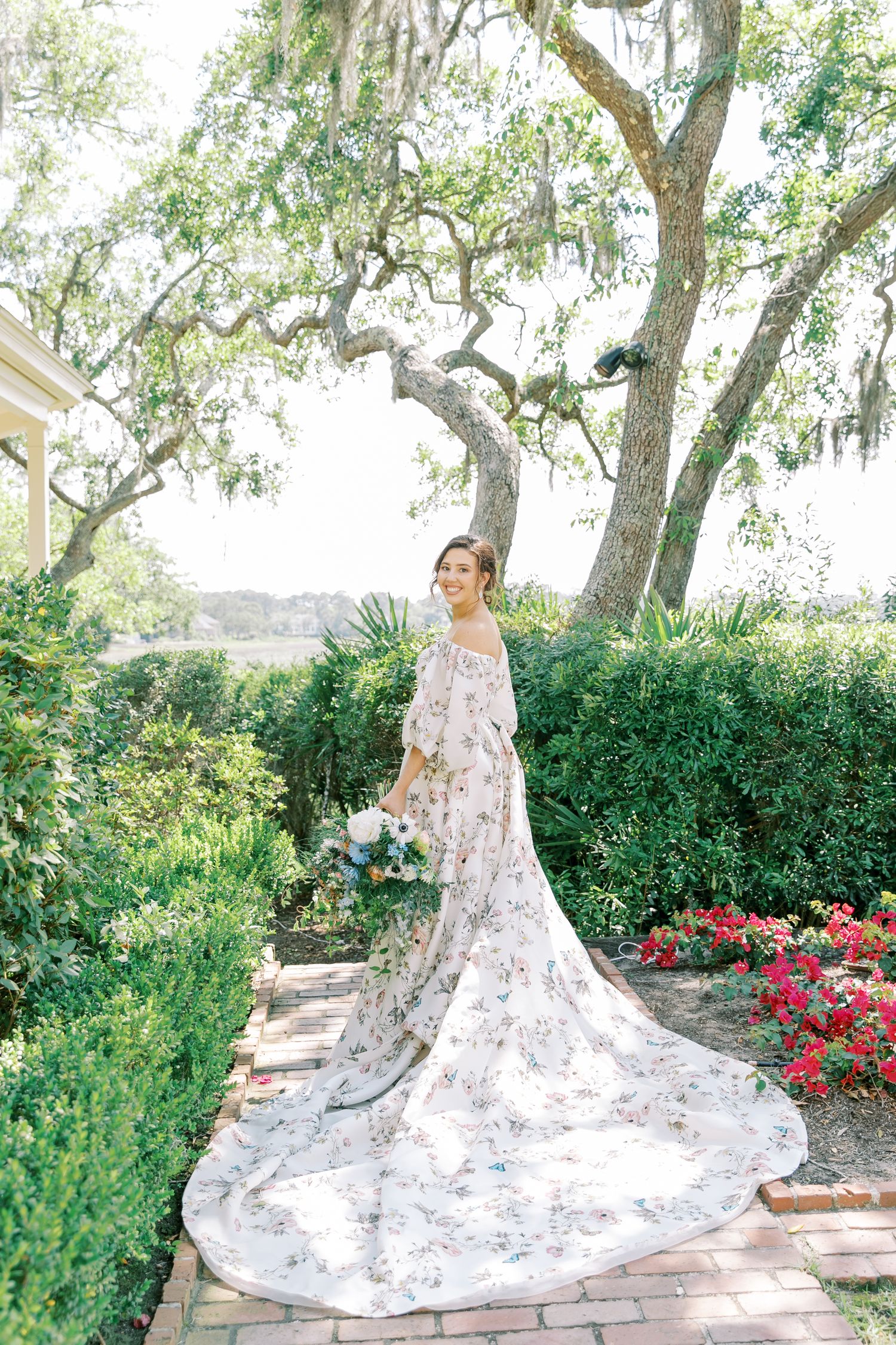 Bride in floral dress with long train poses in garden with Spanish moss trees and red flowers.