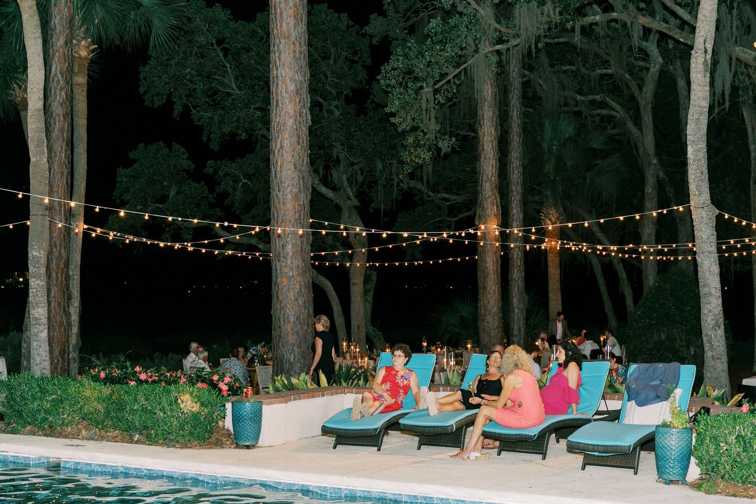 Pool party setting with string lights and lounge chairs under towering pine trees at dusk.