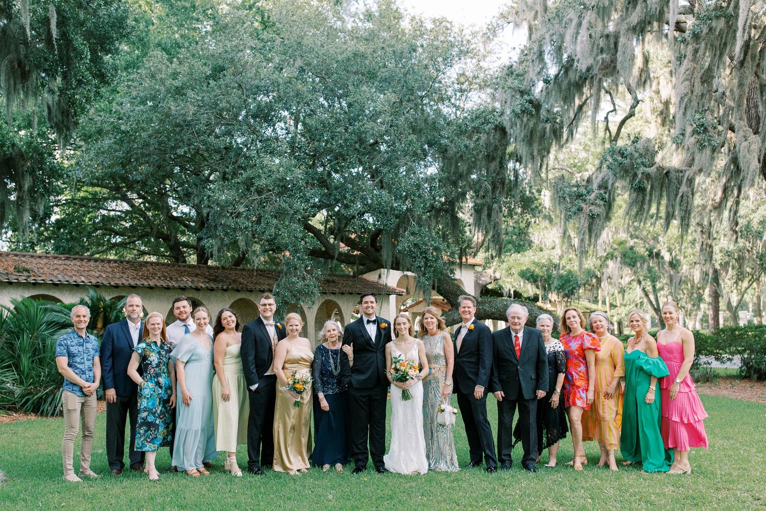 Wedding party members gather for formal group photos under moss-draped oak trees in an outdoor garden setting.