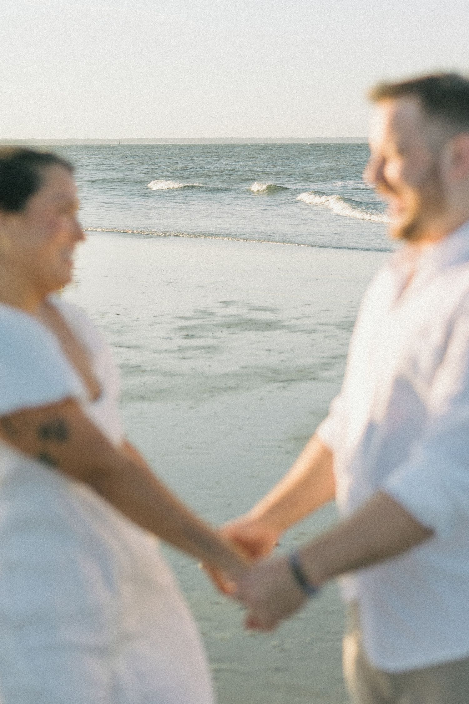 A couple in white outfits holds hands while walking along the beach at sunset.