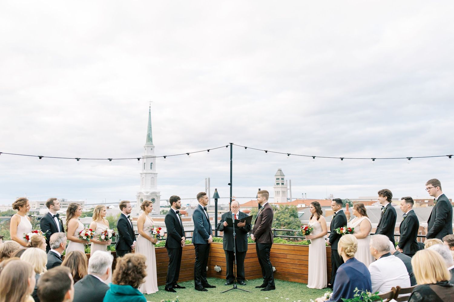 A rooftop wedding ceremony takes place with string lights overhead and a white church steeple visible in the background.