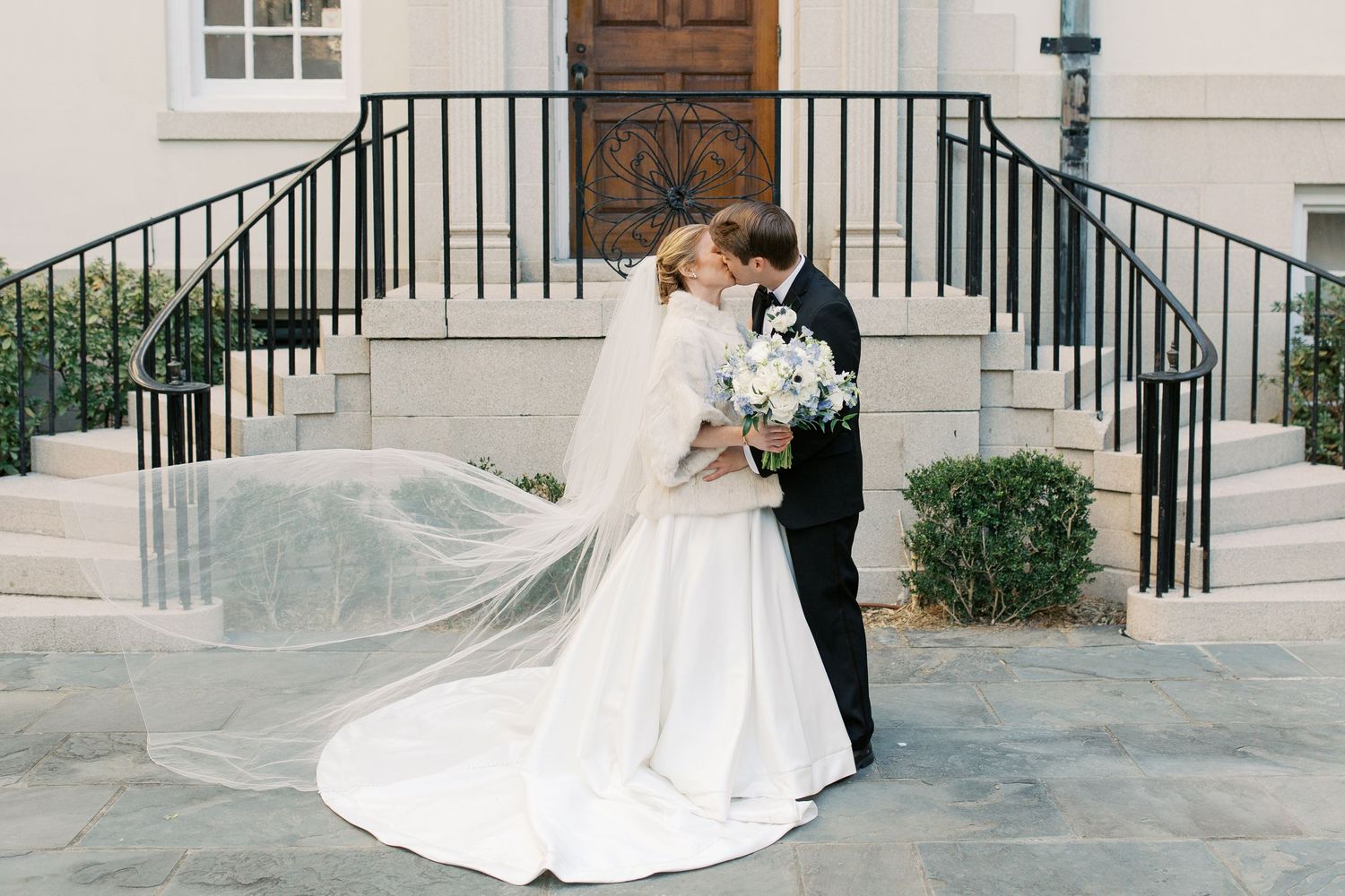 A romantic wedding couple shares intimate moments in front of a white building with black railings and elegant architecture.