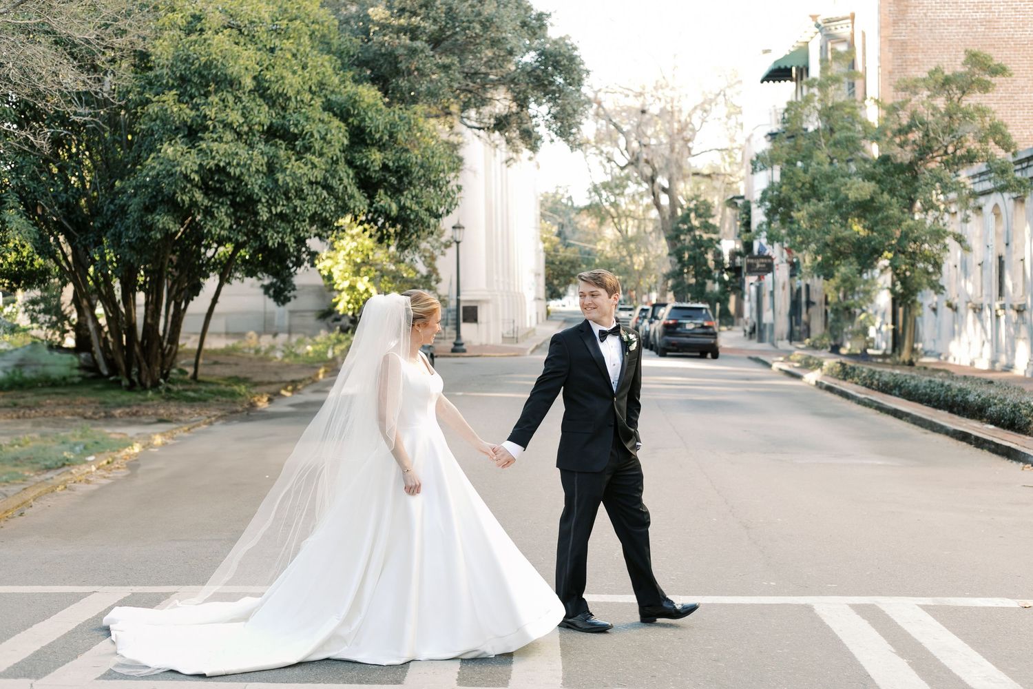 A couple in wedding attire walking across a crosswalk on a tree-lined city street.