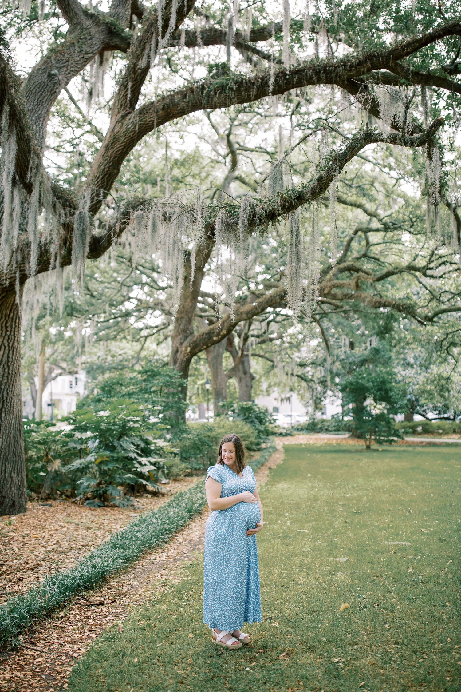 Woman in blue dress poses against scenic backdrop of moss-draped oak trees in a park setting.