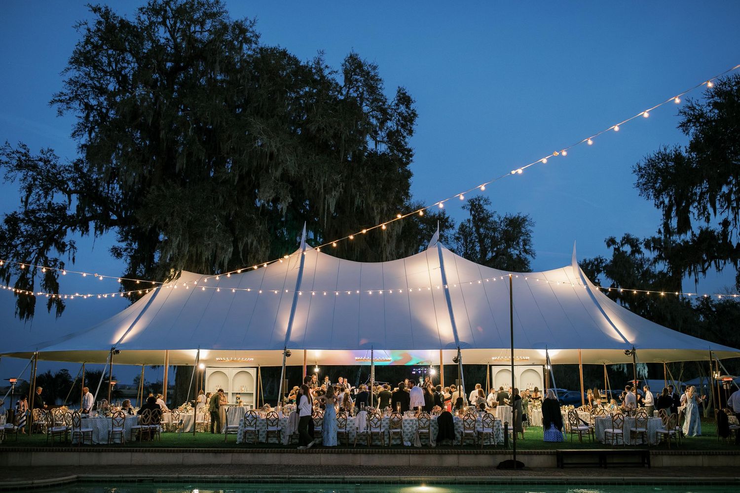 String lights illuminate an outdoor wedding tent and dance floor area against a dusky blue sky.