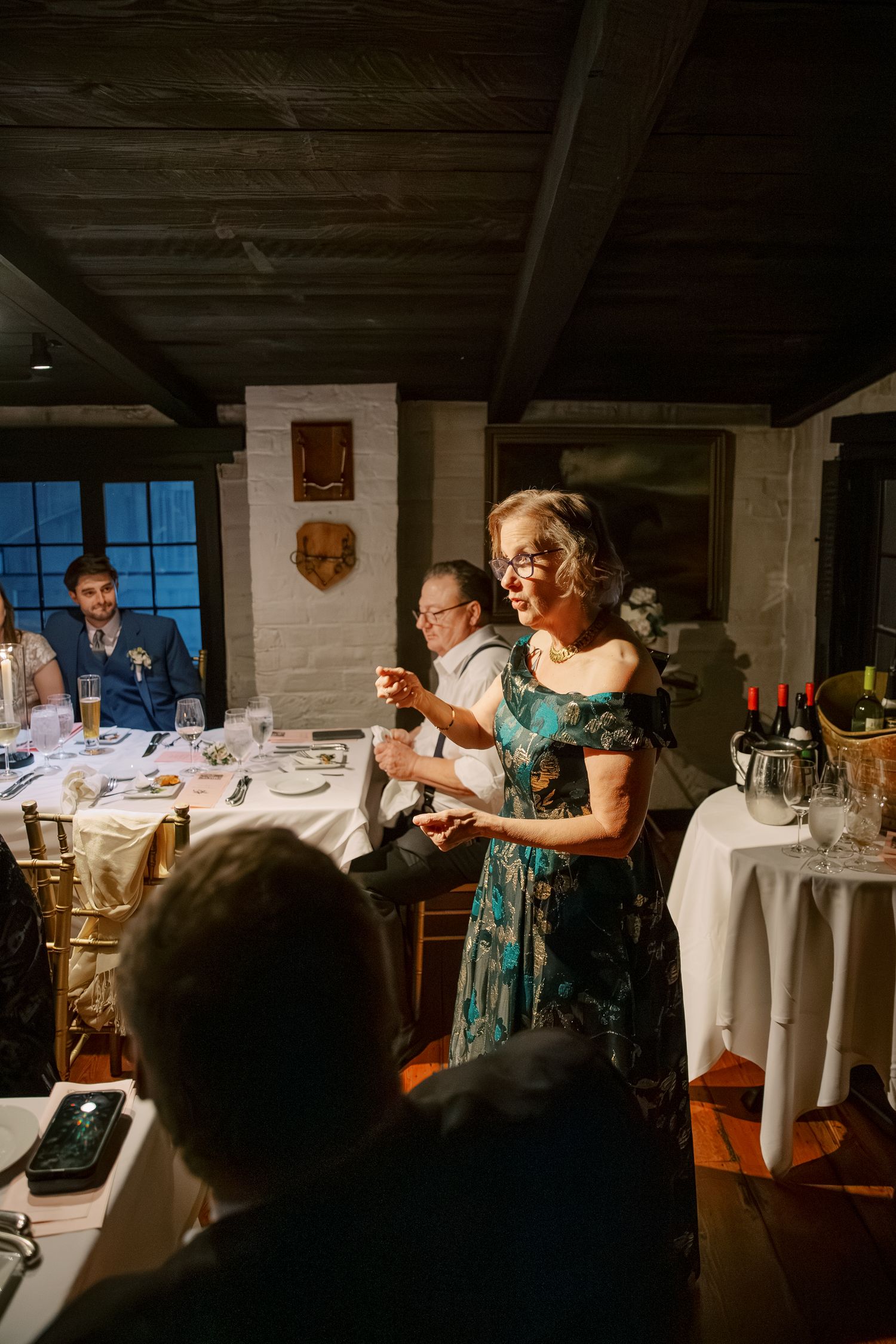 A person in a floral dress speaks to guests at an elegant indoor dinner reception with white tablecloths.