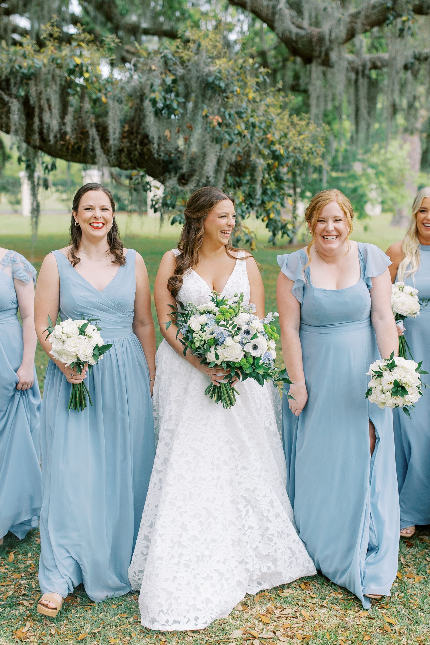 Wedding party in light blue bridesmaid dresses and white bridal gown pose outdoors under Spanish moss draped trees.