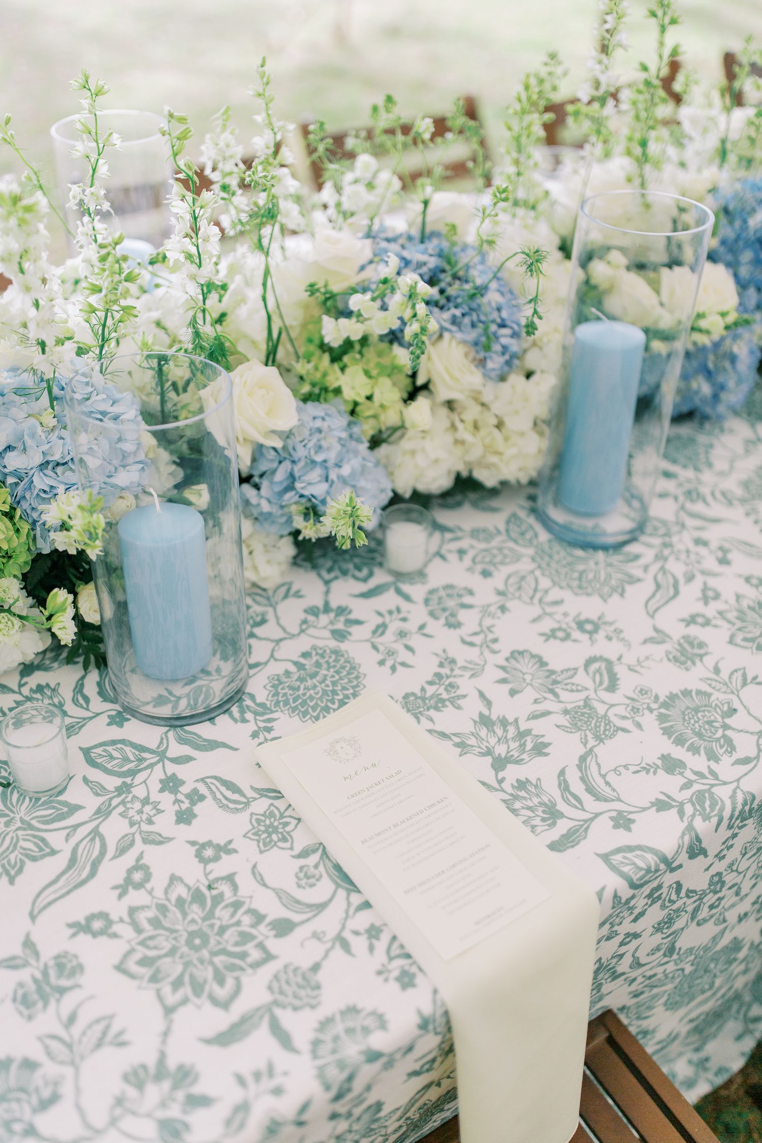 Elegant table setting with blue and white floral centerpieces, candles, and patterned tablecloth.